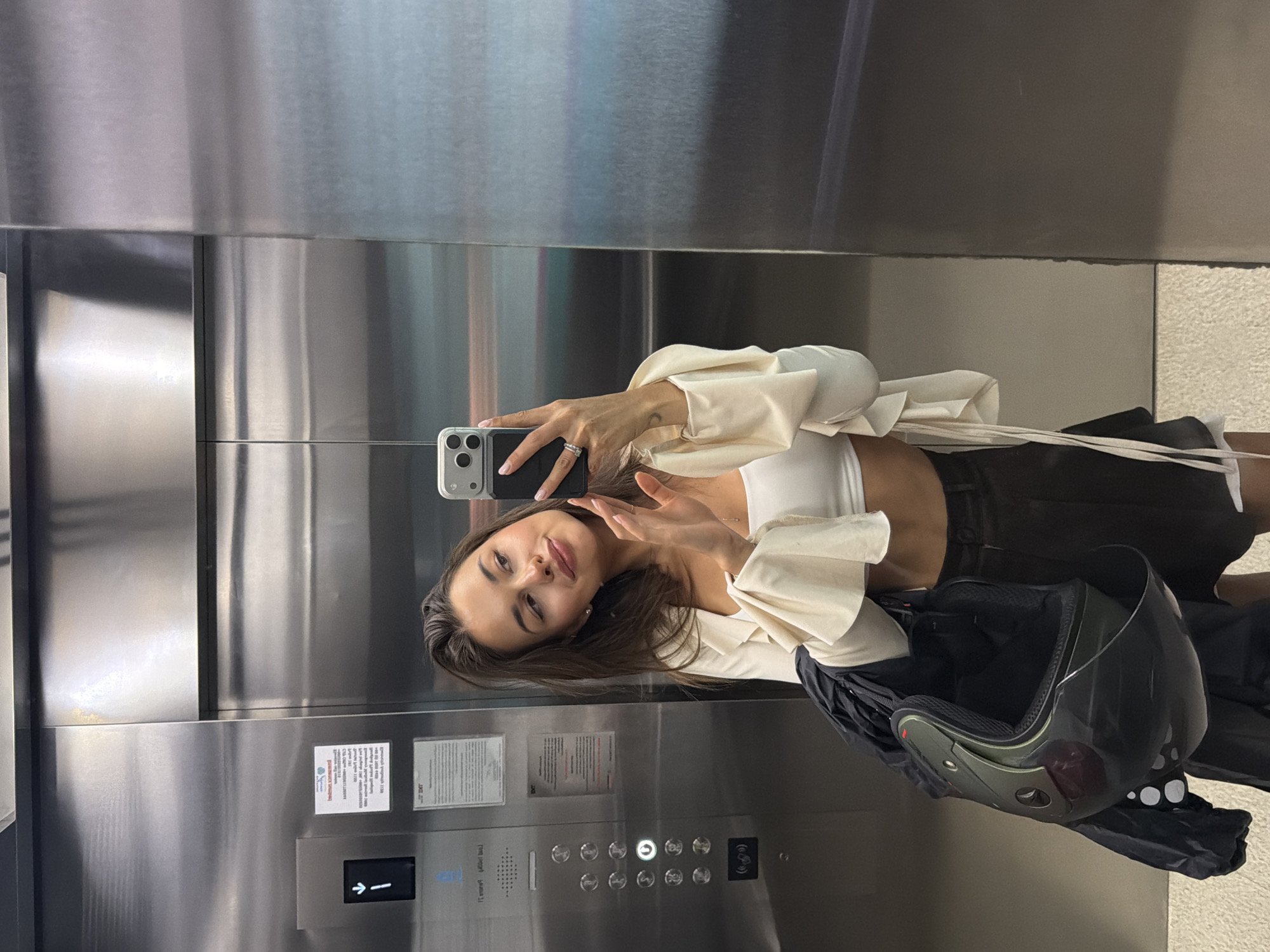 A young woman is taking a selfie in an elevator. She is holding a smartphone in her left hand and posing with her right hand. She is wearing a white top and dark shorts or a skirt. A motorcycle helmet is lying near her. The elevator has stainless steel walls and control panel with buttons and signs. The environment suggests that the image was taken in a modern building, likely in Choeng Thale, Thailand based on the user's prompt.