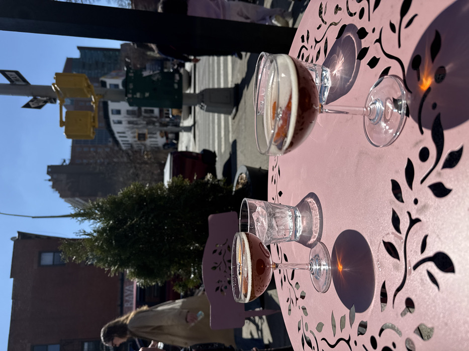 The scene depicts an outdoor patio on a clear, sunny day, located at the intersection of Macdougal Street and Minetta Lane/Waverly Place in Greenwich Village, Manhattan, New York City, United States. The establishment is identified as "Rava" by a visible sign on a brick building in the midground.
In the immediate foreground, a pink metal outdoor table with a laser-cut floral pattern holds two stemmed coupe glasses containing a dark brown liquid with a white foam head and an orange peel garnish. A third, shorter glass filled with ice and clear liquid is positioned between them. Sunlight casts distinct shadows of the glassware and the table's intricate design, with a bright reflection evident on the table's surface. A partial view of a matching pink metal chair is visible on the left.
Beyond the table, a row of green conical evergreen shrubs separates the patio from the sidewalk and street. A woman with long, light brown hair, wearing a light-colored long coat, walks leftward behind the shrubs, holding a light blue object in her hand. A red pickup truck is parked on the street behind the shrubs.
The background reveals a typical New York City street intersection. A street light pole equipped with yellow traffic signals and a "ONE WAY" arrow sign pointing right is prominent. A dark green utility box is affixed to a pole. Multiple multi-story residential and commercial buildings line the thoroughfares. On the far right, two figures, an adult and a child wearing a light purple garment, are observed walking away from the viewer, crossing the street. The sky is bright blue and cloudless.