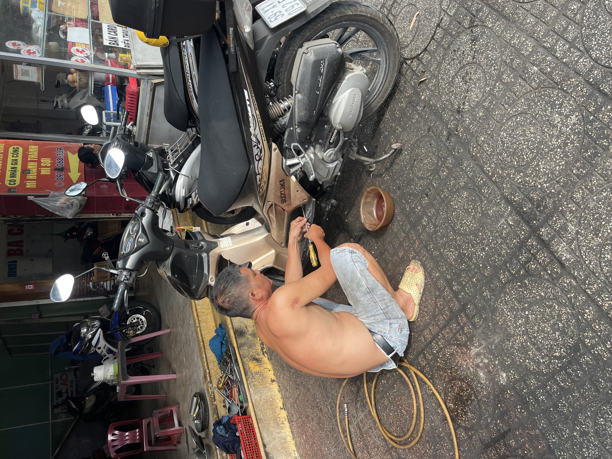 The scene, photographed from a high angle, depicts a shirtless man in light blue jeans and yellow sandals repairing a brown and black Suzuki motorcycle on a patterned pavement. The man is kneeling close to the motorcycle, using a screwdriver. Nearby, a brown bowl and coiled yellow hose sit on the ground. In the background, a motorcycle shop displays other motorcycles and parts, with signage visible, including text in Vietnamese. A storefront is also visible, behind the motorcycle shop. The motorcycle has a license plate partially visible as "59-X1". Based on signage, the location is in Bac Nha Trang Ward, Vietnam.
