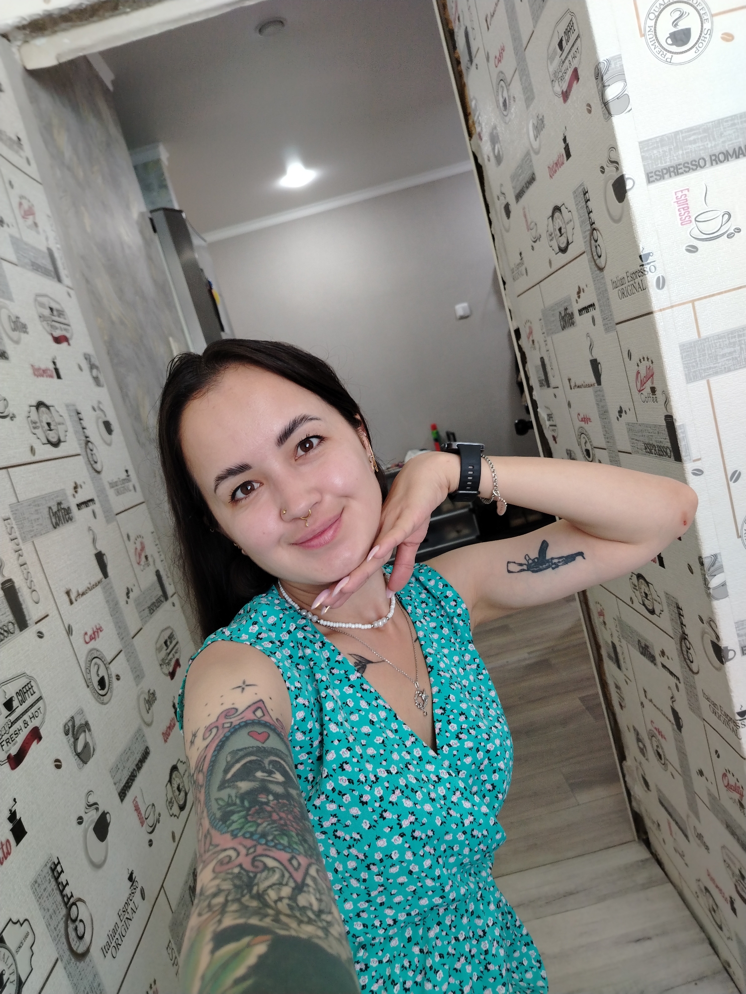 A young woman with dark hair, brown eyes, and a nose ring takes a selfie in what appears to be a narrow hallway. She is wearing a teal floral dress and a pearl necklace. She has tattoos on both arms: one on her right arm depicts a gun, and the other on her left arm is a colorful, intricate design featuring a raccoon. She is smiling and resting her chin on her hand. The walls of the hallway are covered in wallpaper with a coffee-themed pattern. The lighting suggests an indoor setting. There is no visible indication of the specific location within Yanaul, Russia.