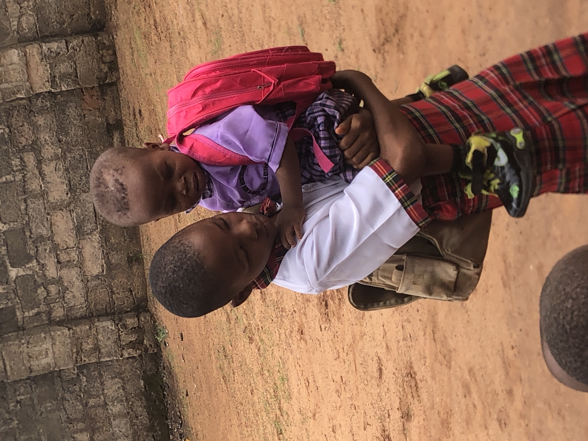 The image captures two children in what appears to be Ukwu-Abwa, Nigeria, engaged in a piggyback ride. An older child, wearing a white short-sleeved shirt with a red and black plaid collar, khaki shorts, and black shoes with green and yellow details, is carrying a younger child on their back. The older child's arms are around the younger one, providing support.
The younger child is dressed in a purple t-shirt and a bright red backpack. Their head rests gently on the older child's back, and they have a calm, almost smiling expression. Visible text on the younger child's purple shirt reads "MONTESSORI SCHOOL EDUCATION," suggesting they attend a Montessori school.
The setting features a reddish-brown dirt ground, with some sparse green weeds visible. On the left side of the frame, there is a textured wall made of greyish-brown stones or bricks. The natural lighting indicates it is daytime. The scene conveys a sense of warmth and companionship between the two children, possibly on their way to or from school, given their attire and the backpack.