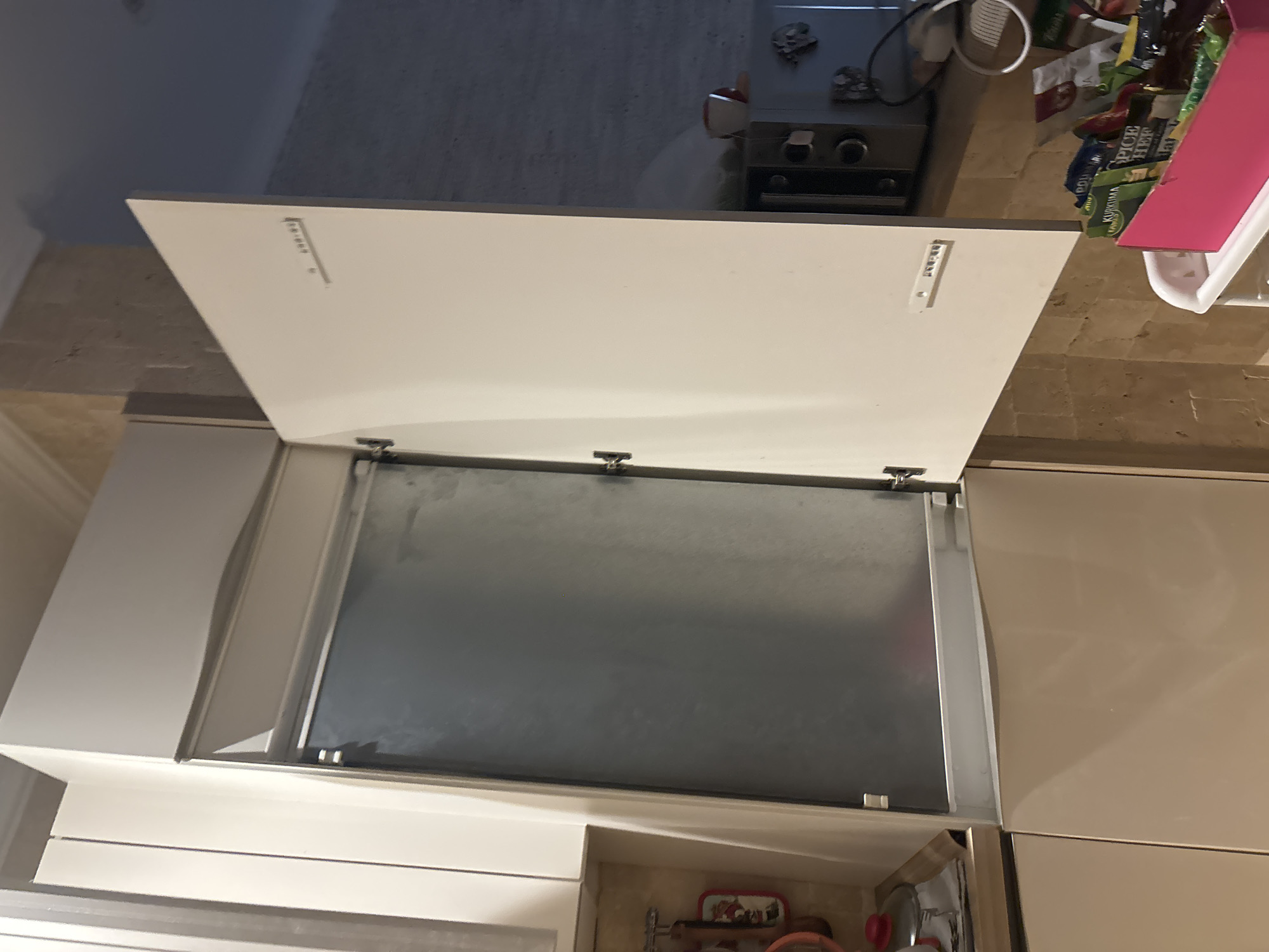 The image shows an interior scene of a kitchen in Budva, Montenegro. The focus is on an open cabinet with a large, metallic interior. One of the cabinet doors is open at a 90-degree angle, revealing the silver interior. The cabinet is light-colored, possibly white or cream. To the left of the open cabinet, another cabinet door is visible, closed. Below the open cabinet are two light-colored drawers or cabinet doors.
On the counter to the right of the open cabinet, a microwave is visible, along with a cup with a tea bag. Further to the right, a bright pink box filled with various packets of spices or food items is placed on a shelf or rolling cart. The wall behind the counter appears to be tiled with a light-colored, textured stone or brick pattern. The ceiling is visible at the top, with decorative molding.