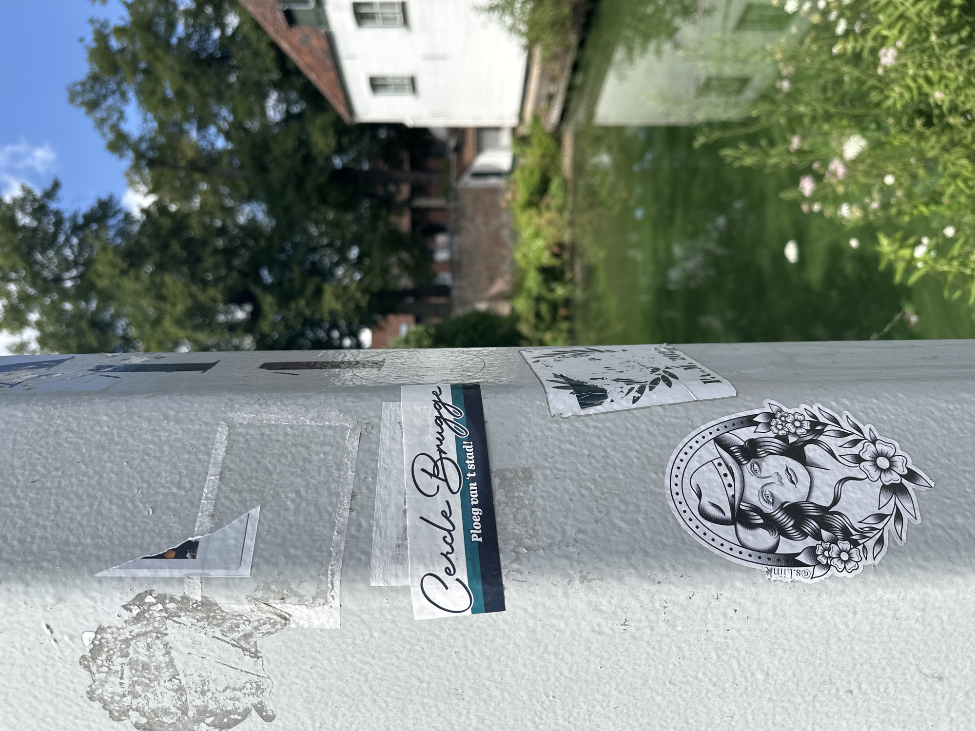 The image shows a close-up view of a railing with several stickers on it. In the background, a canal is visible, reflecting buildings and trees. The water appears green, and the trees are lush and full, suggesting it is a warm day, possibly summer or spring. The sky is blue with some scattered clouds. A white building with windows is prominently featured in the reflection, along with other darker structures and greenery.
On the railing, there are at least three distinct stickers. One is a rectangular sticker with a script font that reads "Cercle Brugge" and below it, "Ploeg van 't stad!". This sticker has blue and green stripes. To the right of this, there is an oval-shaped sticker with a black and white illustration of a woman's face surrounded by flowers and leaves. The woman has long, wavy hair and appears to be depicted in a stylized, almost tattoo-like art style. Below this illustration, faint text is visible, possibly a signature or artist's handle, which seems to be "@s.liink". Further to the left, there are remnants of other stickers or tape, including a torn triangular piece of a sticker that reveals a dark image beneath. The railing itself is painted a light gray and appears to be textured. The overall scene suggests an outdoor setting in a city, likely Bruges, Belgium, given the text on one of the stickers. The sun is shining, casting a bright light on the railing and the stickers.