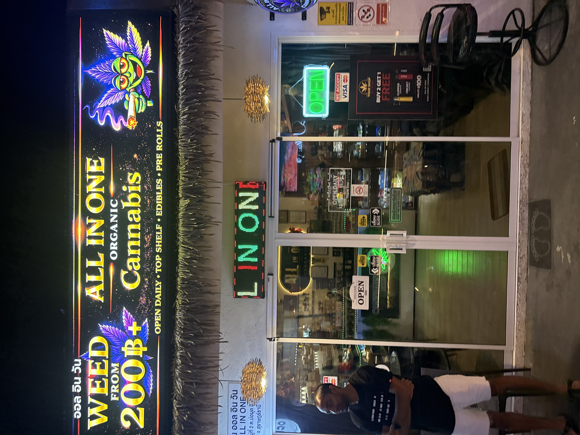 This image shows the exterior of a cannabis shop named "WEED ALL IN ONE ORGANIC CANNABIS" located in Baan Chaweng, Thailand. The shop features a prominent illuminated sign with a cartoonish cannabis leaf character smoking a joint. The entrance is a glass door with several signs, including an "OPEN" neon sign, a "WE ACCEPT VISA" sticker, and a "BUY 2 GET 1 FREE" promotional poster. A man in a black t-shirt and white shorts stands to the left of the entrance. The shop appears to be on a street with a thatched overhang above the entrance.