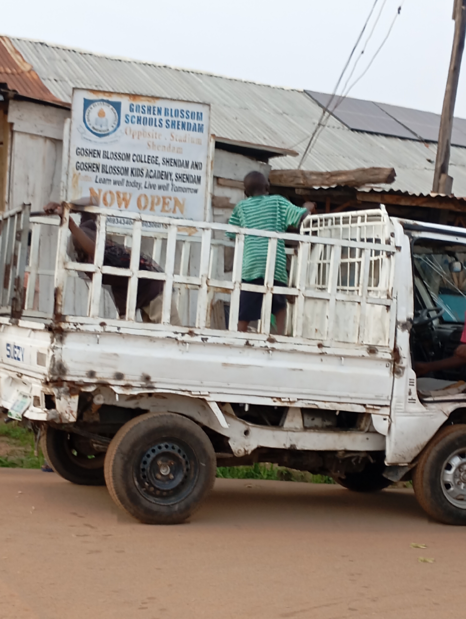 A white truck with a caged bed is parked on a dirt road in Shendam, Nigeria. The truck bed is occupied by several people, some of whom are obscured. In the background, a corrugated metal roof is visible, partially covered by solar panels. A sign on the building reads "GOSHEN BLOSSOM SCHOOLS SHENDAM Opposite Stadium Shendam" and "GOSHEN BLOSSOM COLLEGE, SHENDAM AND GOSHEN BLOSSOM KIDS ACADEMY, SHENDAM Learn well today, Live well Tomorrow NOW OPEN". The truck's body shows signs of rust and damage.