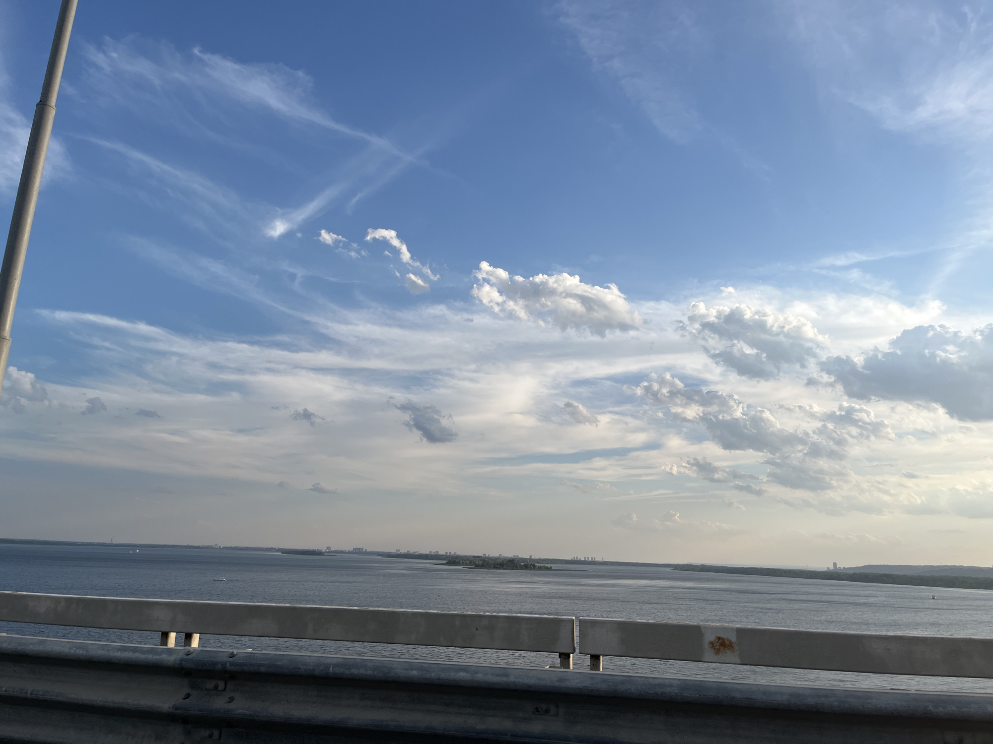 This image was taken from a bridge overlooking a wide body of water, likely a river or a reservoir, with a distant shoreline visible. The sky is a vibrant blue, filled with a mix of wispy cirrus clouds and larger, more defined cumulus clouds, suggesting fair weather with some atmospheric activity. The water is a deep blue-gray and appears calm, with a few small boats or vessels visible on its surface. Along the distant shoreline, there are hints of a city or settlement, with buildings and structures scattered along the landscape. A metal guardrail is prominently in the foreground, indicating the viewpoint is from a road or elevated walkway. The overall impression is one of a scenic, expansive view of a waterway and the urban or semi-urban landscape beyond.