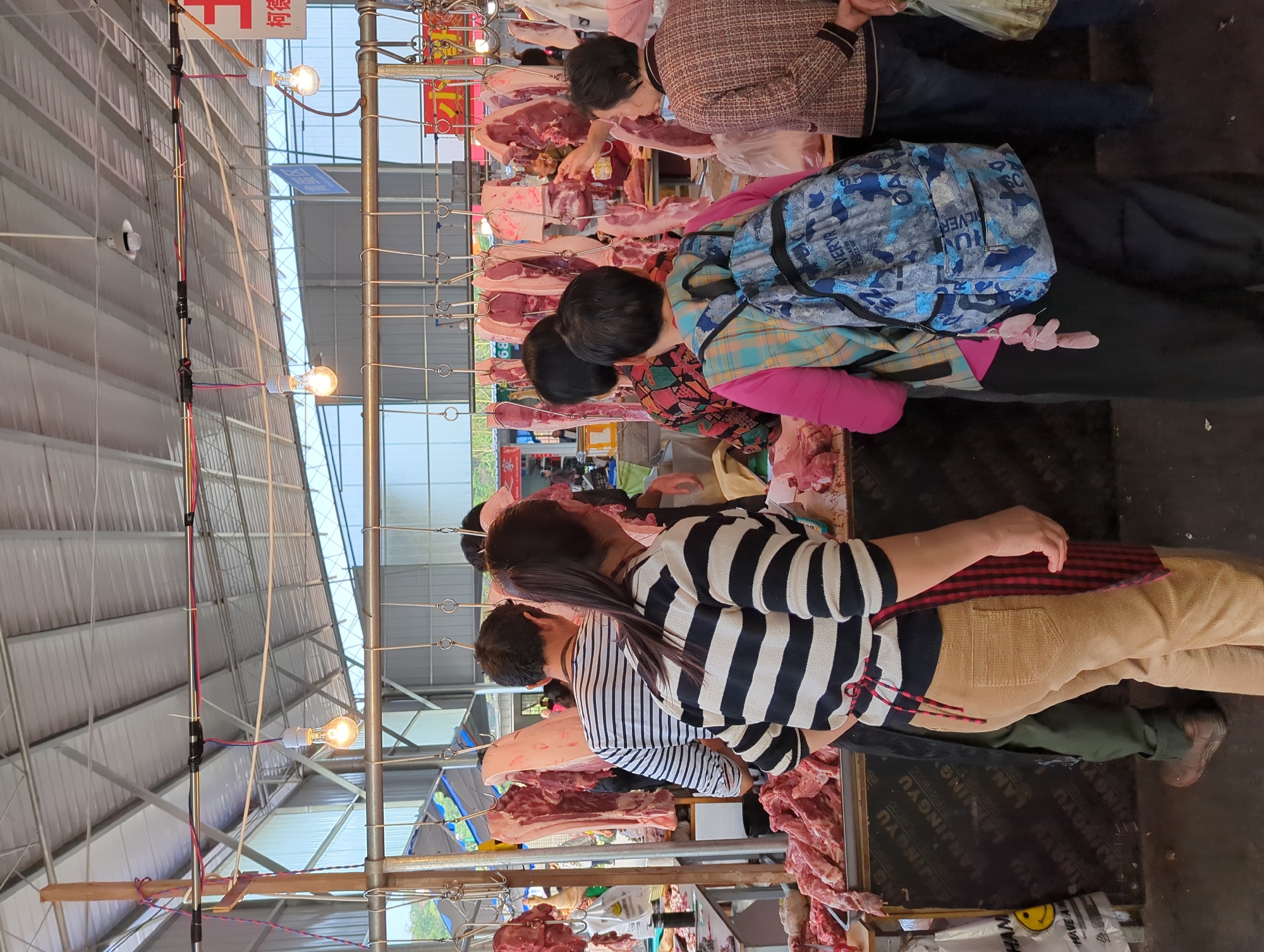 The image displays a bustling market stall, presented sideways, specializing in the sale of raw meat, likely pork. Several people, mostly women, are actively engaged in the scene. In the foreground, a person wearing a black and white striped shirt, tan corduroy pants, and a red and black plaid apron stands next to a display of meat, facing inwards, suggesting they are a vendor or market worker. Further into the stall, another individual wearing a brown tweed-like jacket is seen examining a large cut of meat hanging from metal hooks. Adjacent to them, a person wearing a colorful plaid shirt over a pink top and carrying a blue camouflage backpack is also visible; text on the backpack includes "ALVERYR," "SILVER," "OA," "SD," and "HUNT."
Large cuts of raw meat are prominently displayed, both hanging from hooks on metal structures and laid out on tables or counters. The market space features a high ceiling constructed from corrugated panels. Illumination comes from multiple bare incandescent light bulbs strung along wires, casting a warm light over the stall. Various texts are discernible within the image: a red sign with Chinese characters, including "柯德" (Kēdé), and repeated text on a dark surface on the floor or a low platform that reads "ANJINGYU," "LANJINGYU," "PLYWOOD," and "HAVE A." The overall scene depicts a busy, functional market environment during what appears to be daytime.