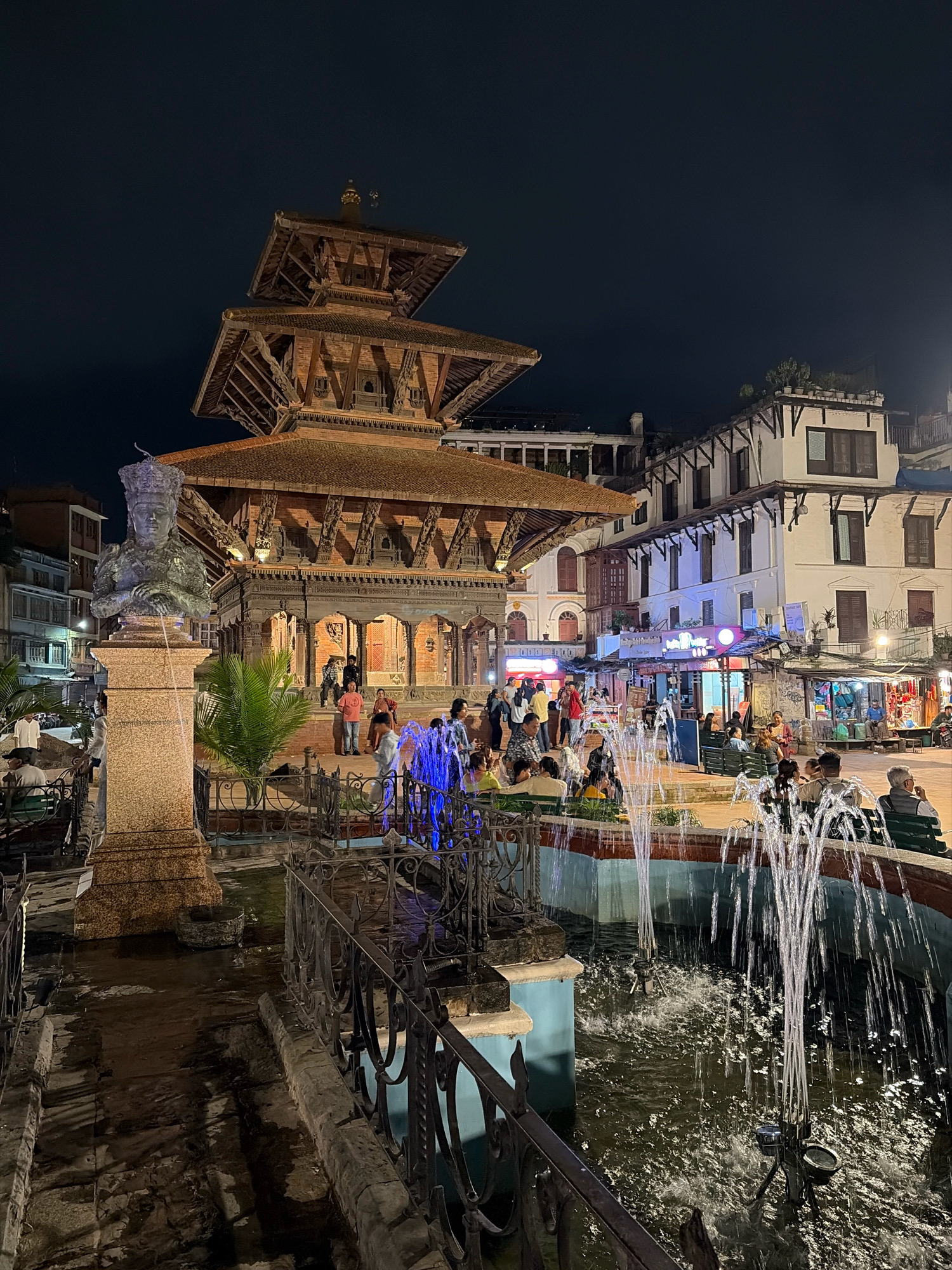 The image captures a vibrant nighttime scene in Patan Durbar Square, Lalitpur, Nepal. In the foreground, a fountain with multiple jets of water illuminated by blue and white lights is active. A dark metal fence separates the viewer from the fountain. To the left of the fountain is a stone pedestal topped with a bronze bust of a regal figure, adorned with a crown and jewelry, which also appears to be a water feature. Beyond the fountain and statue, a large, multi-tiered pagoda-style temple made of dark wood and brick dominates the background, its intricate carvings and architectural details visible under the night sky. Several people are gathered in front of the temple, some standing and others sitting. To the right of the temple, a street scene unfolds with several buildings lining the square. These buildings feature white facades, dark windows, and balconies. Shops and stalls are visible at street level, with illuminated signs and goods displayed, suggesting a bustling marketplace. People are walking and congregating in this area as well, enjoying the evening atmosphere. The overall scene is lively, with artificial lighting from the fountain and the shops creating a dynamic and engaging ambiance in this historic square.