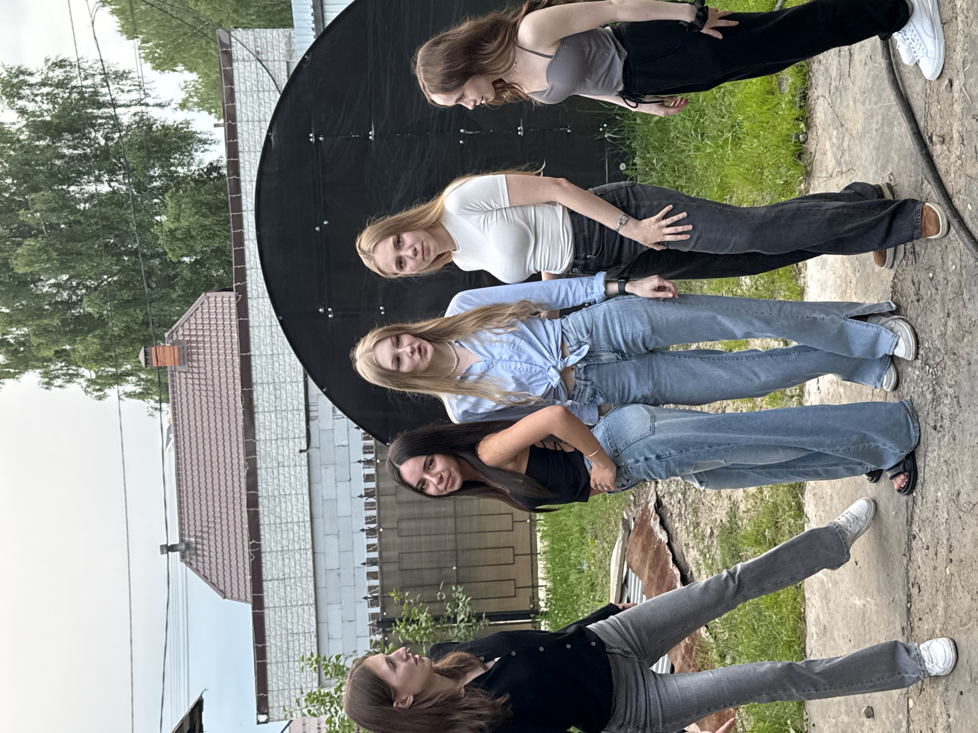 Five young women are gathered outdoors in what appears to be a backyard or residential area. The woman on the far left, wearing a black cardigan and grey jeans, is facing left and looking forward, with her left leg extended forward. The second woman from the left, in a black tank top and light blue jeans, is facing towards the camera with her right hand on her hip and a slight smile. The third woman from the left, in a light blue button-down shirt tied at the waist and light blue jeans, is facing towards the camera and slightly to the left, with her hands clasped in front of her. The fourth woman, wearing a white t-shirt and dark grey jeans, is facing the camera with her hands at her sides. The woman on the far right, wearing a grey tank top and black pants, is facing to the left and looking towards the other women. In the background, there is a black arched structure, a fence, and buildings with reddish-brown roofs, consistent with residential architecture. The ground is a mix of dirt and pavement. The overall scene suggests a casual gathering. The presence of the specific architectural style of the buildings and the overall aesthetic are consistent with residential areas in the city of Perm, Russia.
