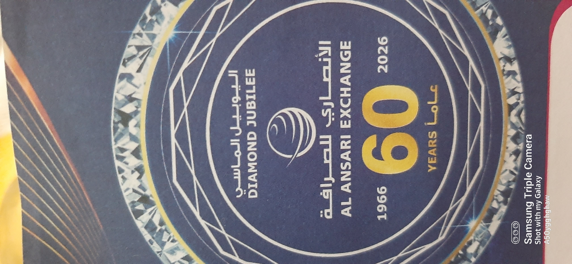 The image displays a close-up view of a circular emblem with a dark blue background. The emblem features several concentric rings and stylized diamond patterns. In the center, a stylized globe symbol is present. Above this, text in Arabic reads "اليوبيل الماسي" (Al Yobel Al Masi) and in English "DIAMOND JUBILEE". Below the globe, Arabic text reads "الأنصاري للصرافة" (Al Ansari Lil Sarrafa), followed by the English text "AL ANSARI EXCHANGE". Further down, the years "1966" and "2026" are shown flanking a large, golden numeral "60". Beneath the numeral "60", the word "YEARS" and Arabic text "عاماً" (Aaman) are visible. The bottom of the image shows the text "Samsung Triple Camera Shot with my Galaxy A50yggghghaw". The overall design suggests a commemorative event for Al Ansari Exchange. There is no indication of the city of Dubai or the United Arab Emirates within the image itself.