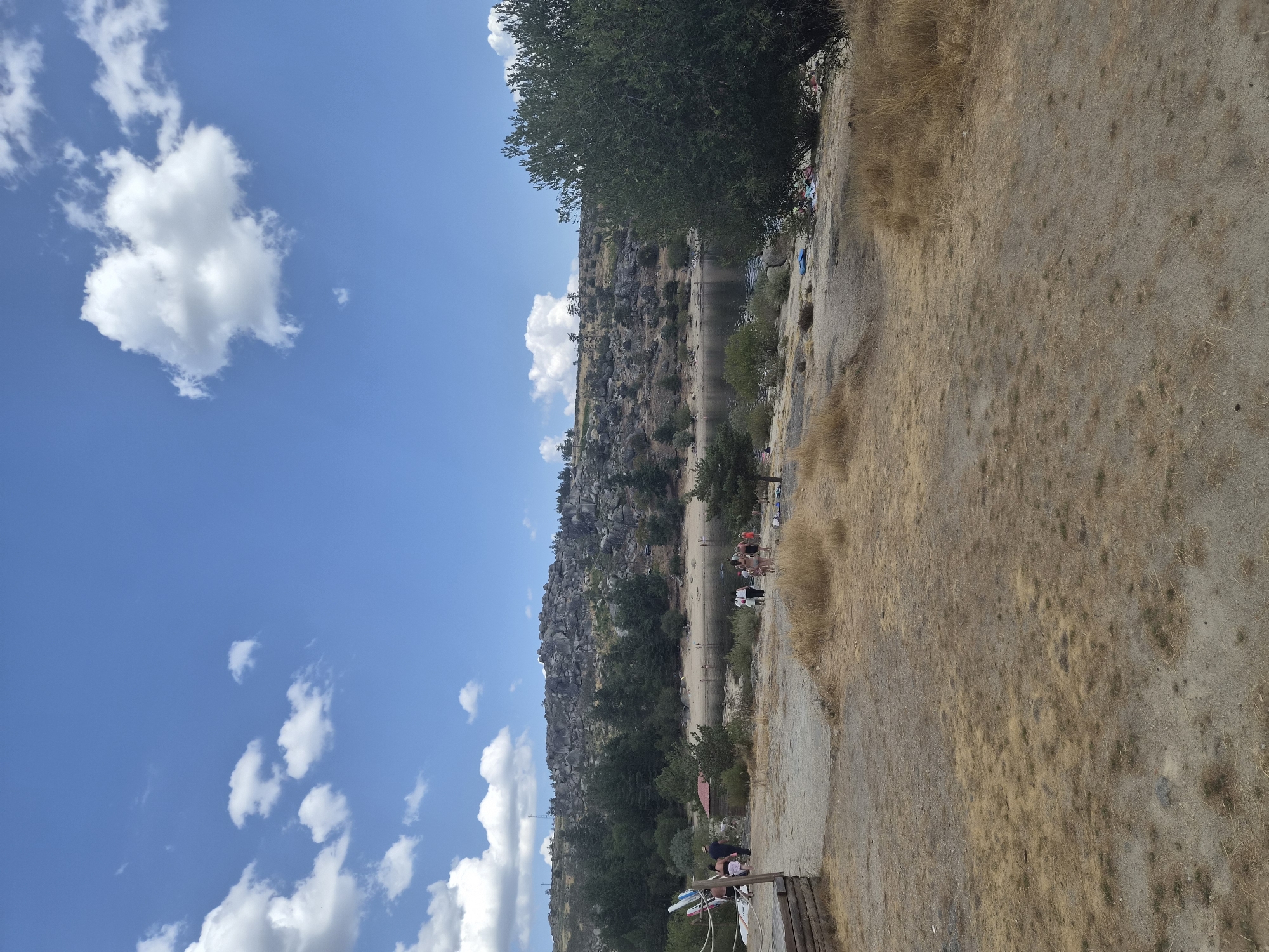 The image depicts a scenic view of a lake or reservoir area, likely in the municipality of Gouveia, Portugal, known for its natural landscapes. A clear blue sky with scattered white cumulus clouds forms the upper portion of the scene. Below the sky, a rugged, rocky hill covered with sparse trees and vegetation slopes down towards the water.
In the foreground, dry, yellowish grass covers a sandy, uneven terrain. To the left, a wooden platform with a railing is visible, and several individuals are gathered nearby, some holding surfboards or paddleboards. Further into the midground, a small red-roofed structure sits amongst trees. Along the edge of the water, a sandy beach is populated by several people, some swimming or relaxing. A large, leafy green tree dominates the right side of the image, partially obscuring the background. The overall impression is that of a recreational area during a sunny day.