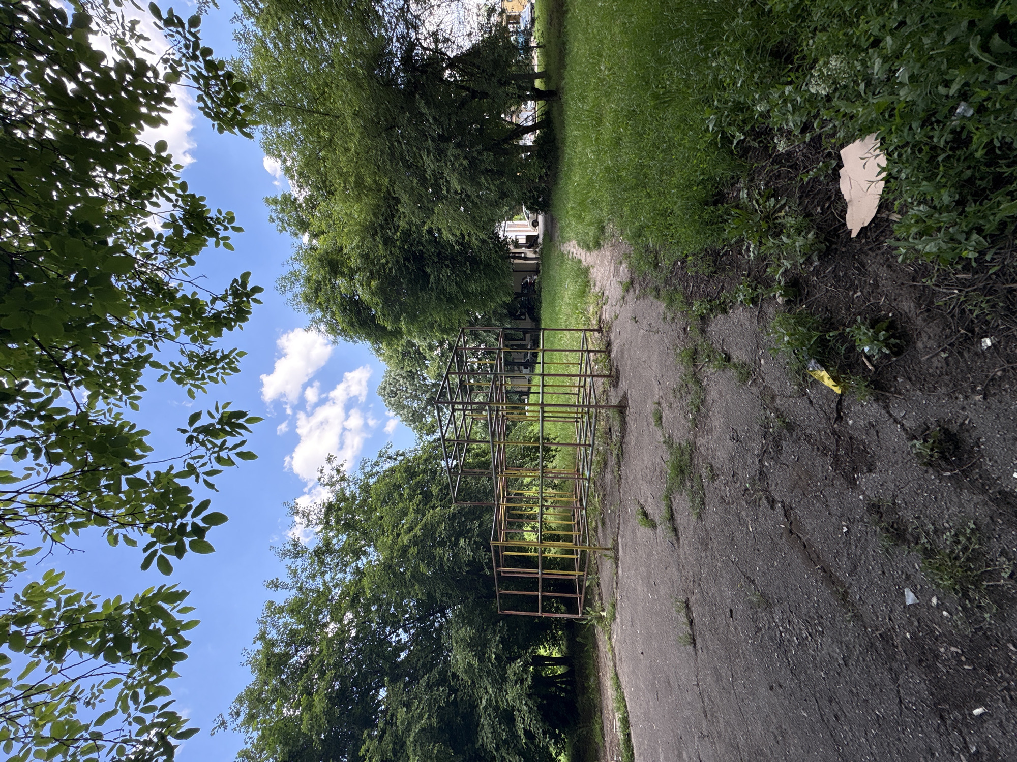 An outdoor scene depicts a concrete pathway leading through a park or wooded area. On the left side of the path, a climbing structure made of metal bars sits beside trees. The path is bordered by grass and vegetation. A piece of trash is visible in the grass. The sky is visible through gaps in the trees and is blue with some clouds. The photo is tilted slightly, giving a skewed perspective. There are no people visible. The location is possibly in the city of Sofia, Bulgaria.