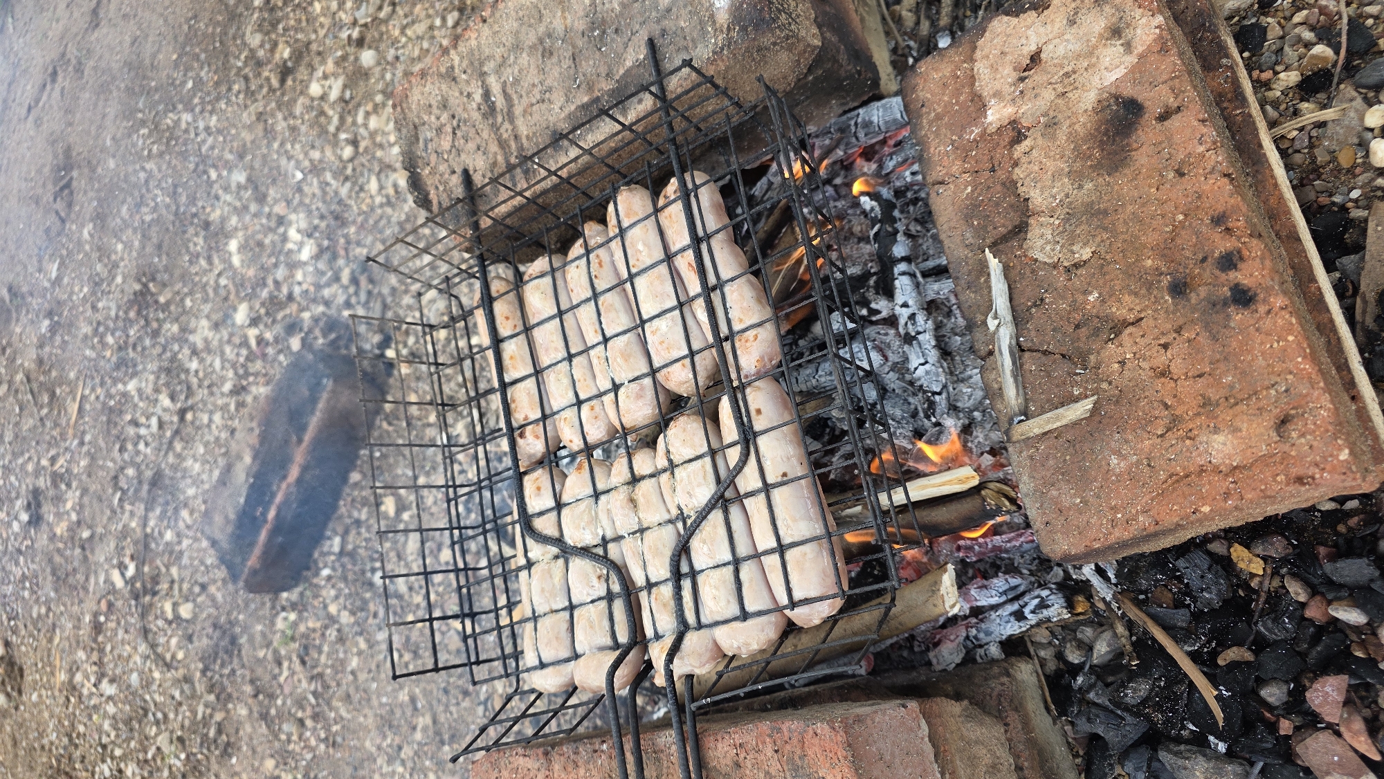 A close-up, high-angle shot shows sausages cooking on a barbecue grill over a fire in what appears to be an outdoor, possibly rural or semi-rural setting, given the gravel and dirt ground. The grill is a black wire rack, holding at least twelve uncooked sausages arranged in rows. The sausages are pale pink and plump. The grill rests on a makeshift structure of red bricks and burning wood, with visible coals and flames underneath. Smoke rises from the barbecue. In the background, a dark, rectangular object, possibly a brick or piece of wood, lies on the gravelly ground. The overall scene depicts food preparation outdoors. The location is specified as Дарьино, Russia.