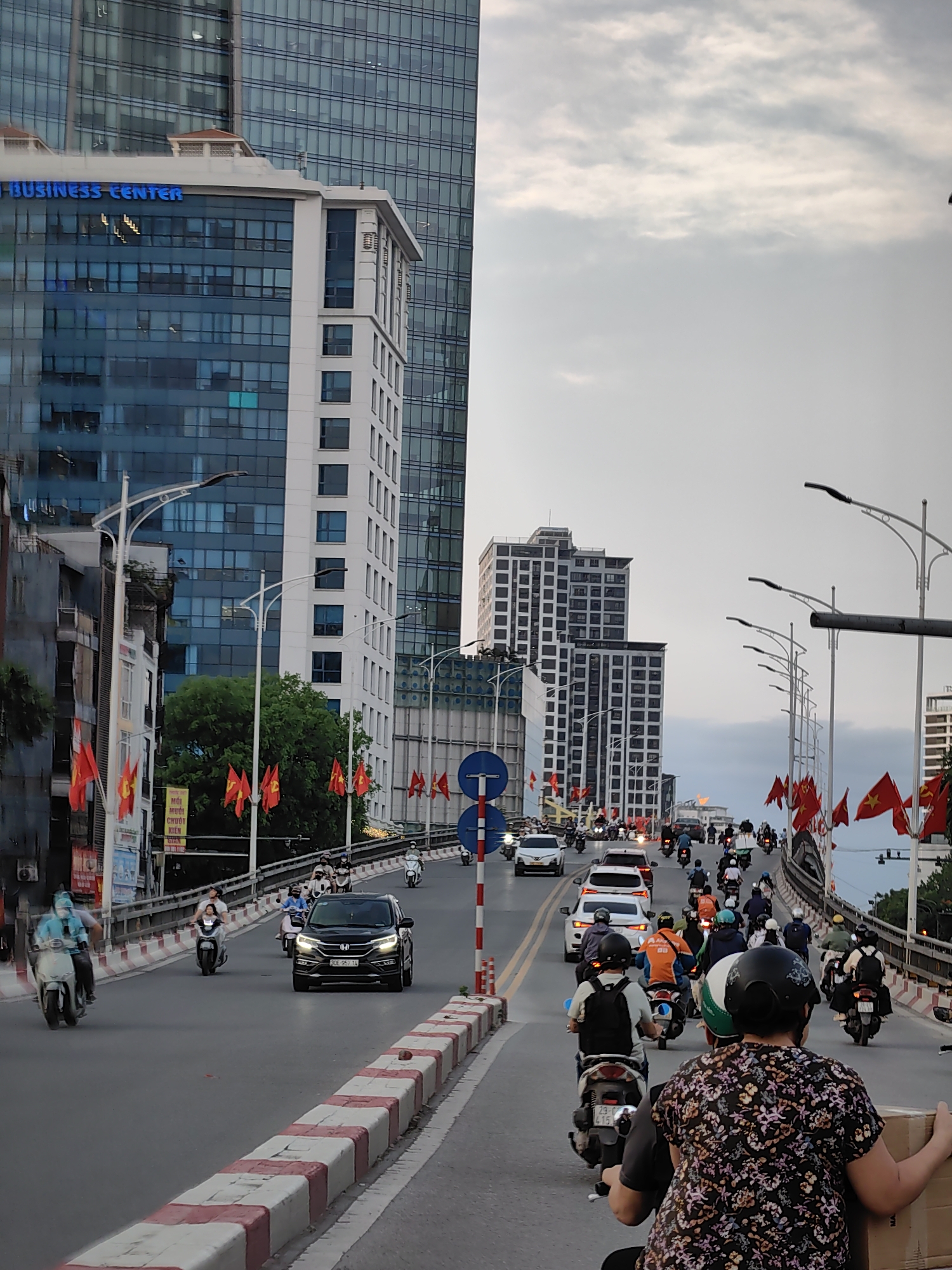 The image captures a bustling urban street or bridge in Hà Nội, Vietnam, likely during the late afternoon or early evening under an overcast or partly cloudy sky. The scene is dominated by a mix of modern and older buildings, with heavy traffic moving along a multi-lane road.
In the foreground and middle ground, numerous motorcycles and scooters, many with riders wearing helmets, are seen in motion. Several cars are also present, including a black SUV in the left lane with the license plate "29G-957.11" and white sedans further back. Red and white striped concrete barriers line the road, separating traffic lanes and the sidewalk/shoulder.
On the left side of the image, a large, modern glass skyscraper stands tall, with a smaller, white building in front of it that has the blue text "BUSINESS CENTER" emblazoned on its facade. Below this, older, multi-story buildings line the street. A row of red Vietnamese flags with yellow stars is prominently displayed along the road's edge and on lampposts, stretching into the distance on both sides of the bridge. One banner on the left shows partially visible Vietnamese text: "NÓI MỚI CHUỘT KIỆN GIAN".
The right side of the image shows more streetlights curving along the bridge. In the background, additional tall, residential-style buildings with numerous windows are visible under the muted sky. The overall impression is one of a dynamic and densely populated city environment, characteristic of Hà Nội.