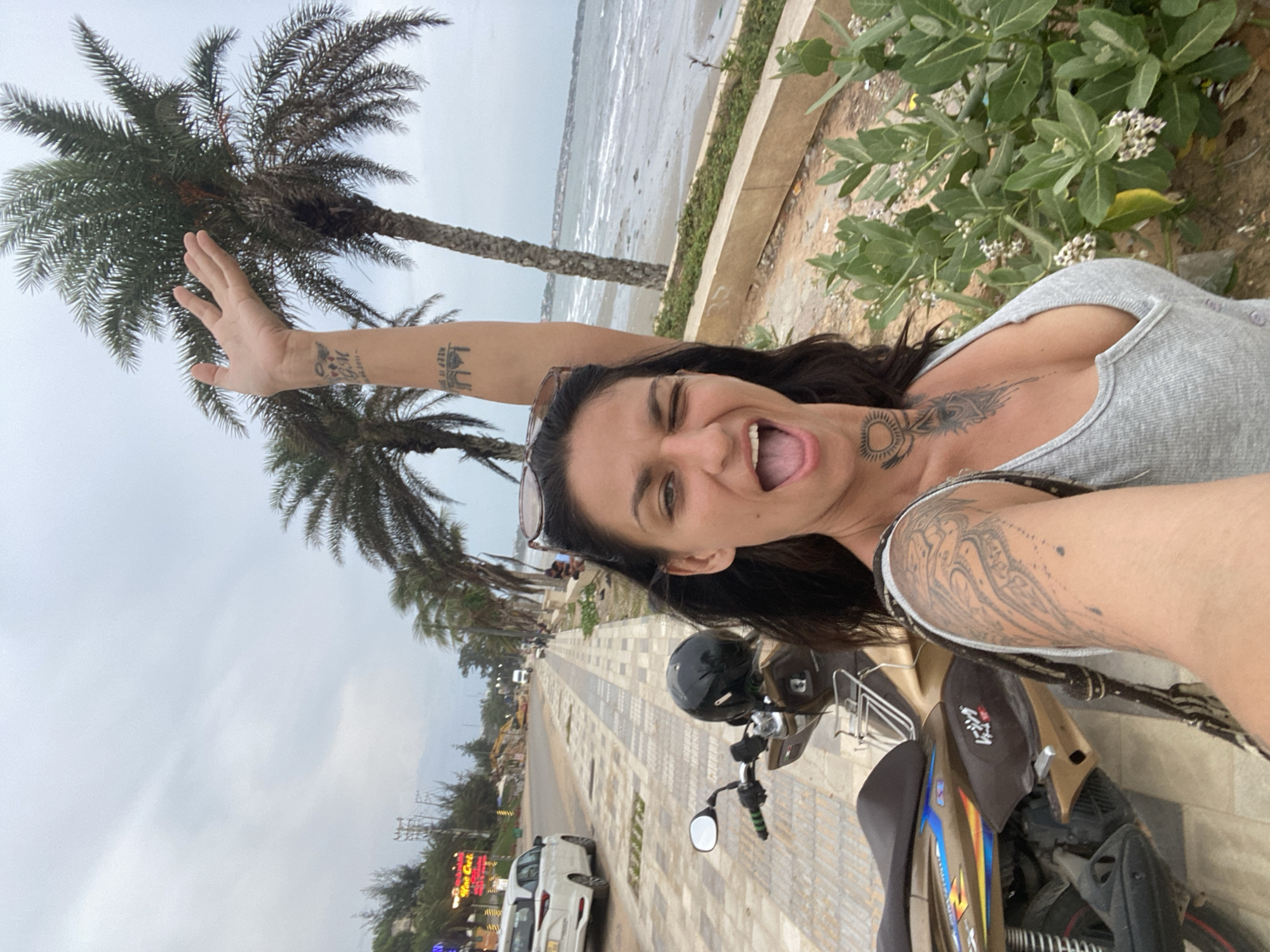 A woman with dark hair and tattoos on her arms, neck, and chest is taking a selfie on a beachfront promenade in Mũi Né, Vietnam. She is wearing a grey tank top and has her right arm raised, waving at the camera. She is winking and her mouth is open in a smile. To her left, a gold and brown scooter is parked. Behind her, palm trees line the promenade, which overlooks a beach and the ocean. The sky is overcast. In the background, there are buildings, a road with cars, and construction.