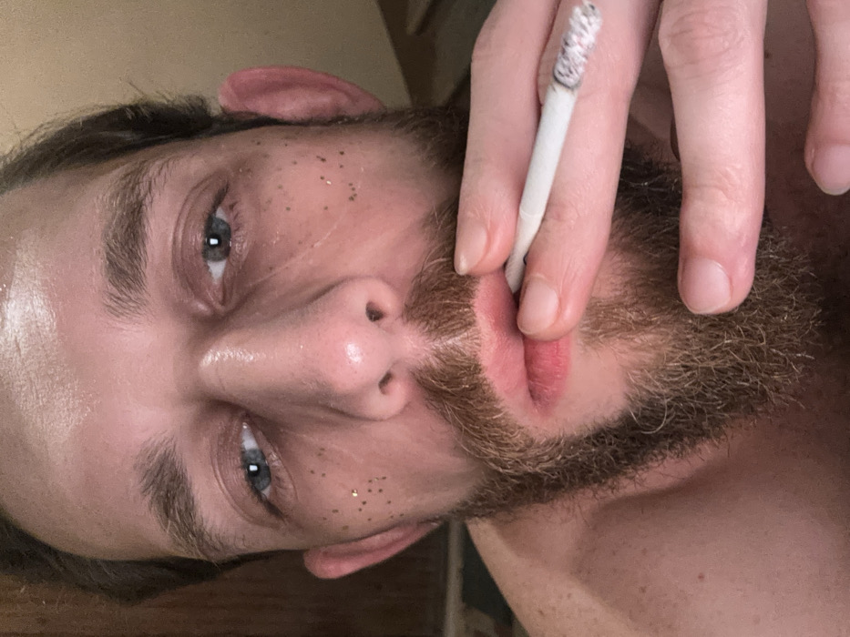 A close-up shot from below showcases a man with light skin, blue eyes, and a light brown beard. Glitter dots adorn his face. He holds a lit cigarette between his fingers, positioned near his lips. The cigarette is partially burned with visible ash. The image gives the impression of being taken indoors with ambient lighting.