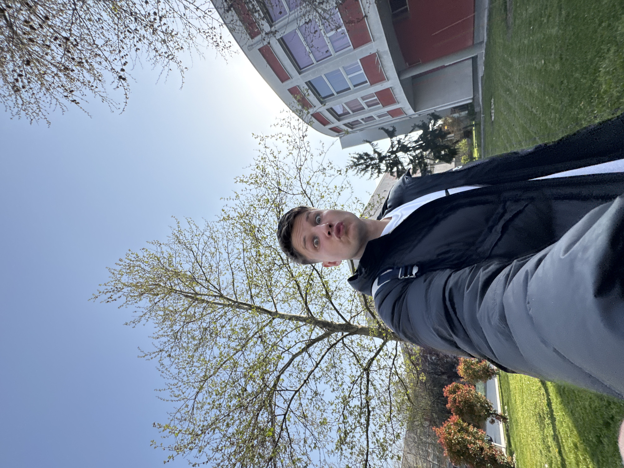 A low-angle, selfie-style photograph depicts a fair-skinned male making a "fish lips" expression, with his head oriented towards the upper-left corner. He is wearing a black jacket over a white garment. Behind him, a tree with budding green leaves fills a large portion of the frame, obscuring the bright blue sky. In the upper portion, the facade of a multi-story building with red and white elements and multiple windows is visible. Green grass is visible on the right side of the image. Based on the architectural style of the building, the location is likely near the University of Food Technologies in Plovdiv, Bulgaria.