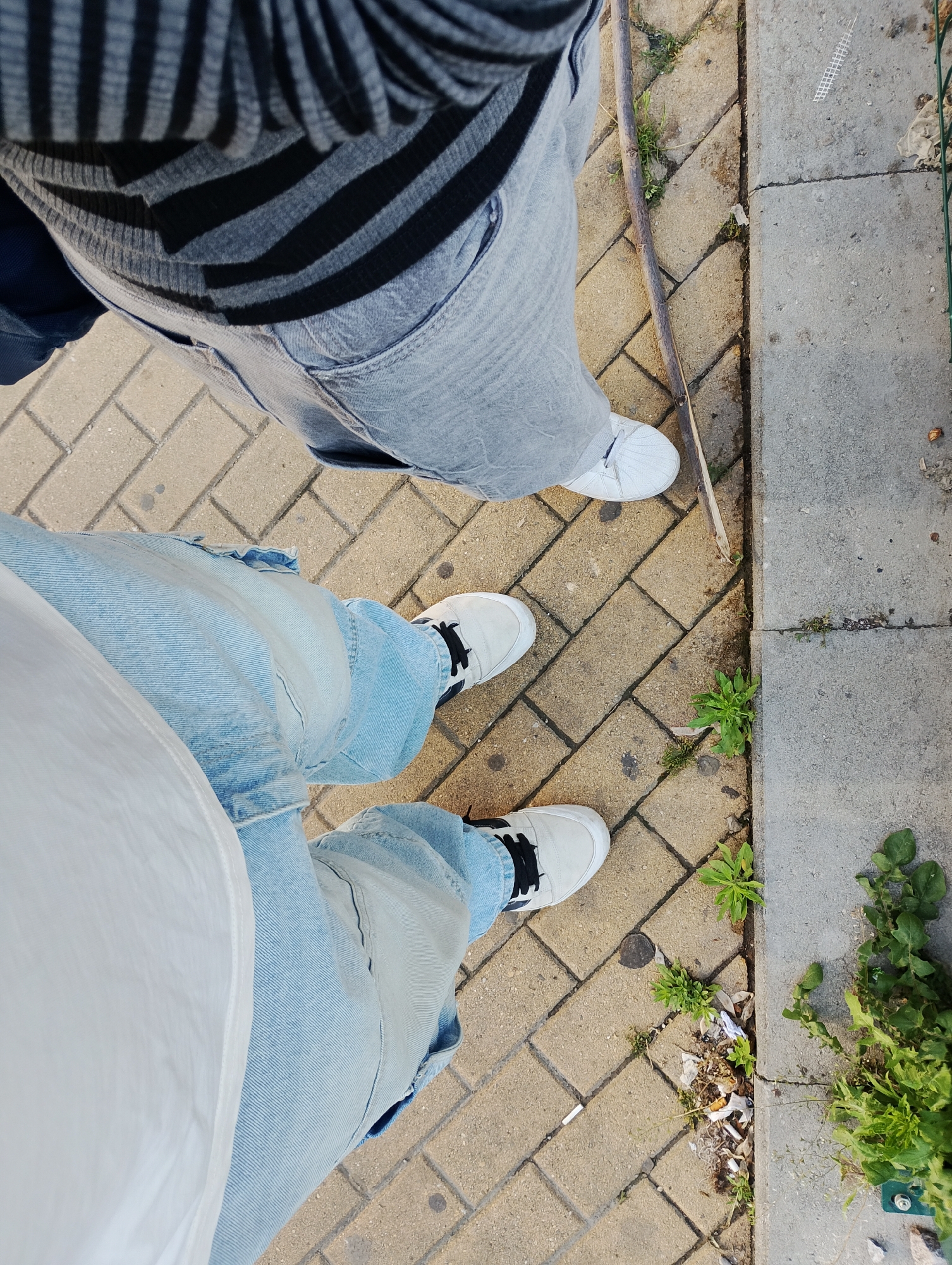 The image, captured from a top-down perspective, shows the lower bodies and feet of two individuals standing on a paved outdoor surface.
The person in the foreground, primarily on the left side of the frame, wears light blue denim jeans that appear somewhat baggy or relaxed fit, paired with a white t-shirt. Their feet are encased in white sneakers featuring black laces.
The second individual, positioned towards the upper part and right side of the frame, wears gray denim jeans and a long-sleeved shirt with alternating gray and black horizontal stripes. Their footwear consists of white sneakers with white laces.
The ground is composed of two distinct sections. The central and left portion features rectangular, yellowish-brown interlocking pavers. To the right, there is a lighter gray concrete slab, possibly part of a sidewalk or curb.
A long, thin wooden stick or branch lies diagonally across the pavers. Small patches of green weeds and plants grow in the cracks between the pavers and along the seam where the pavers meet the concrete slab. Some litter, including what appears to be a discarded cigarette butt and small pieces of paper or plastic, is visible among the vegetation on the paved side.
The lighting indicates a bright, sunny day, with visible shadows cast by the subjects and objects on the ground. No text is discernible in the image.