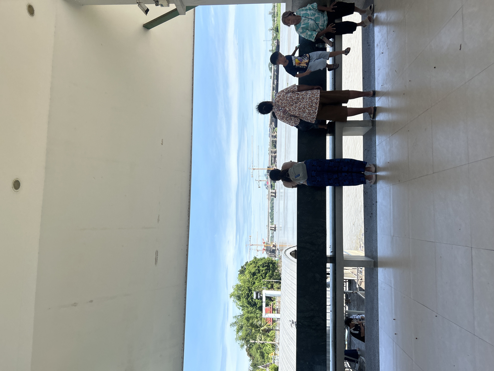 The image depicts a view from an elevated, covered walkway overlooking a river. In the foreground, a white tiled floor is visible, with scattered dark specks. Beyond the walkway's edge, a dark metal railing runs horizontally. Several people are positioned along this railing. On the left, a person wearing blue patterned pants and a grey backpack stands facing away from the camera. Next to them, a person in a patterned shirt and brown shorts stands facing the river. To the right of this person, a young boy in a dark blue t-shirt and light blue shorts is standing and looking down. Further to the right, an elderly woman in a light blue and white patterned shirt sits facing the river. In the background, the river is wide and calm, with a large bridge under construction crossing it. Construction cranes are visible on both sides of the river. Lush green trees line the far bank of the river, and the sky is bright blue with scattered clouds. A CCTV camera is mounted on a support beam in the upper right corner of the image. The setting appears to be a public area, possibly a ferry terminal or a scenic viewpoint in Thai Ko Subdistrict, Thailand.