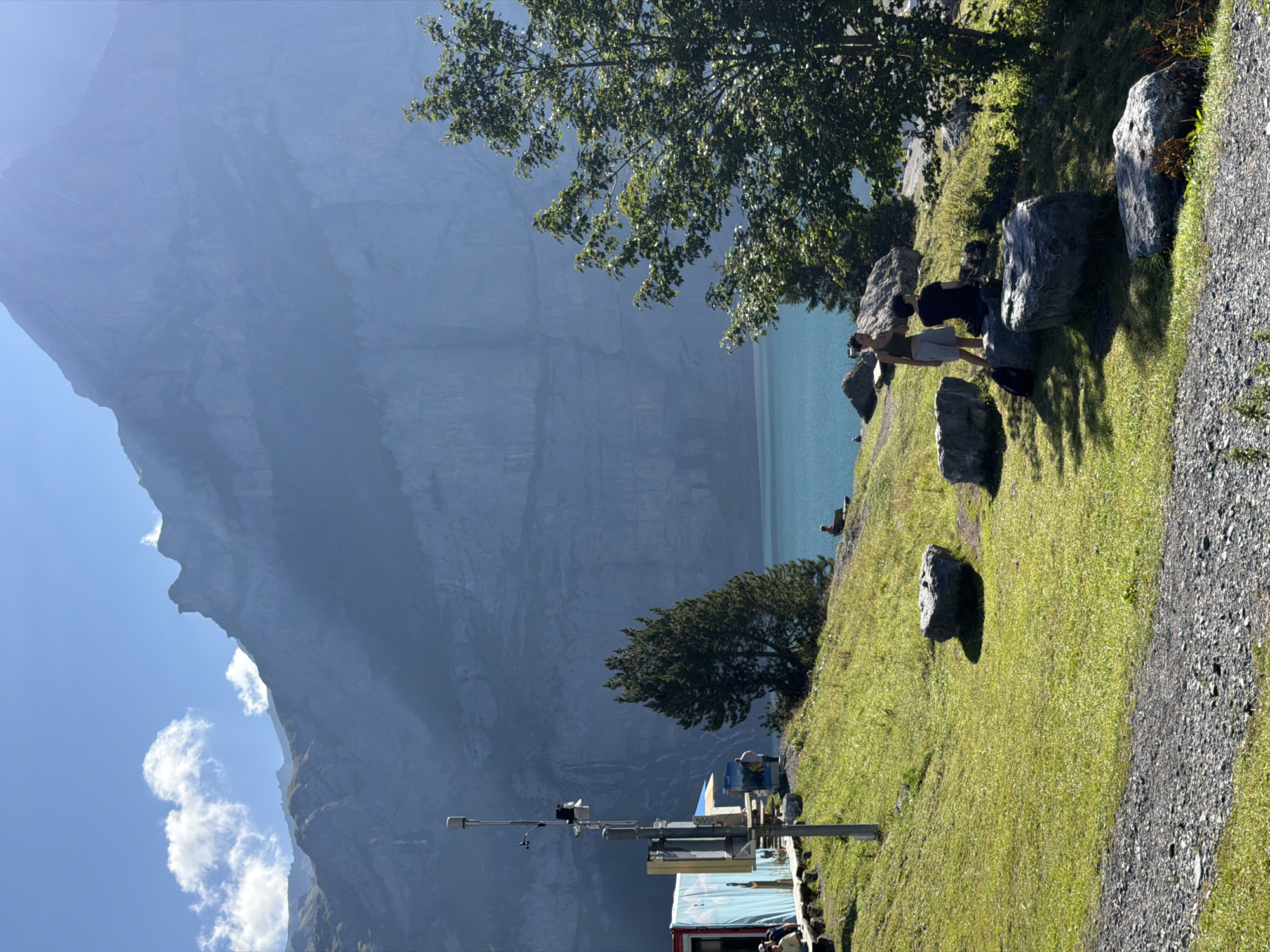 The image depicts a scenic view of a mountain lake in Kandersteg, Switzerland, with a focus on the Oeschinensee lake and its surrounding cliffs. The foreground features a grassy slope with large rocks scattered across it. Two young adults are seated on one of the rocks, facing away from the camera, and appear to be conversing. To the left of the frame, a building with blue and white siding is visible, along with a prominent metal pole topped with various antennas and equipment. Several people are gathered near the building. In the middle ground, the vibrant turquoise water of the lake stretches out, bordered by a lush green hillside and dark green trees. A lone figure is seated on a bench on the hillside, looking out at the lake. The background is dominated by a colossal, sheer mountain face, its rocky texture softened by a blue haze, with a waterfall visible cascading down its side. The sky is a clear blue with a few wispy clouds.