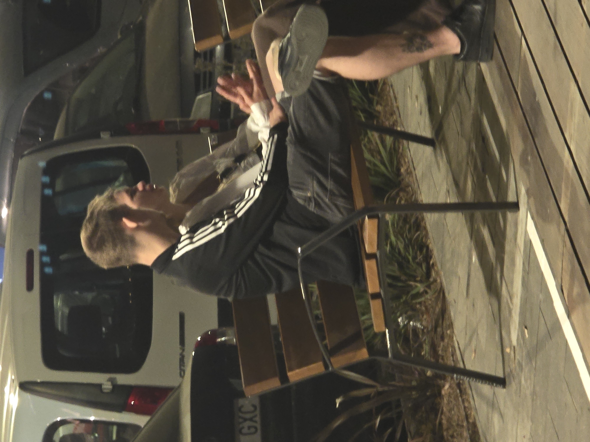 The image shows a man sitting on a wooden chair outdoors, possibly in a public space. He is wearing a black Adidas tracksuit with white stripes, grey shorts, and black shoes on one foot, with the other foot showing a grey shoe. There is a tattoo visible on his leg. He appears to be interacting with someone or something beyond the frame, as he has one hand raised. A white van is parked closely behind him, with part of its license plate ("GXC") visible. The setting suggests an urban environment, specifically in Cornellà de Llobregat, Spain, indicated by the license plate format. The image is taken at night, or in dim lighting conditions.