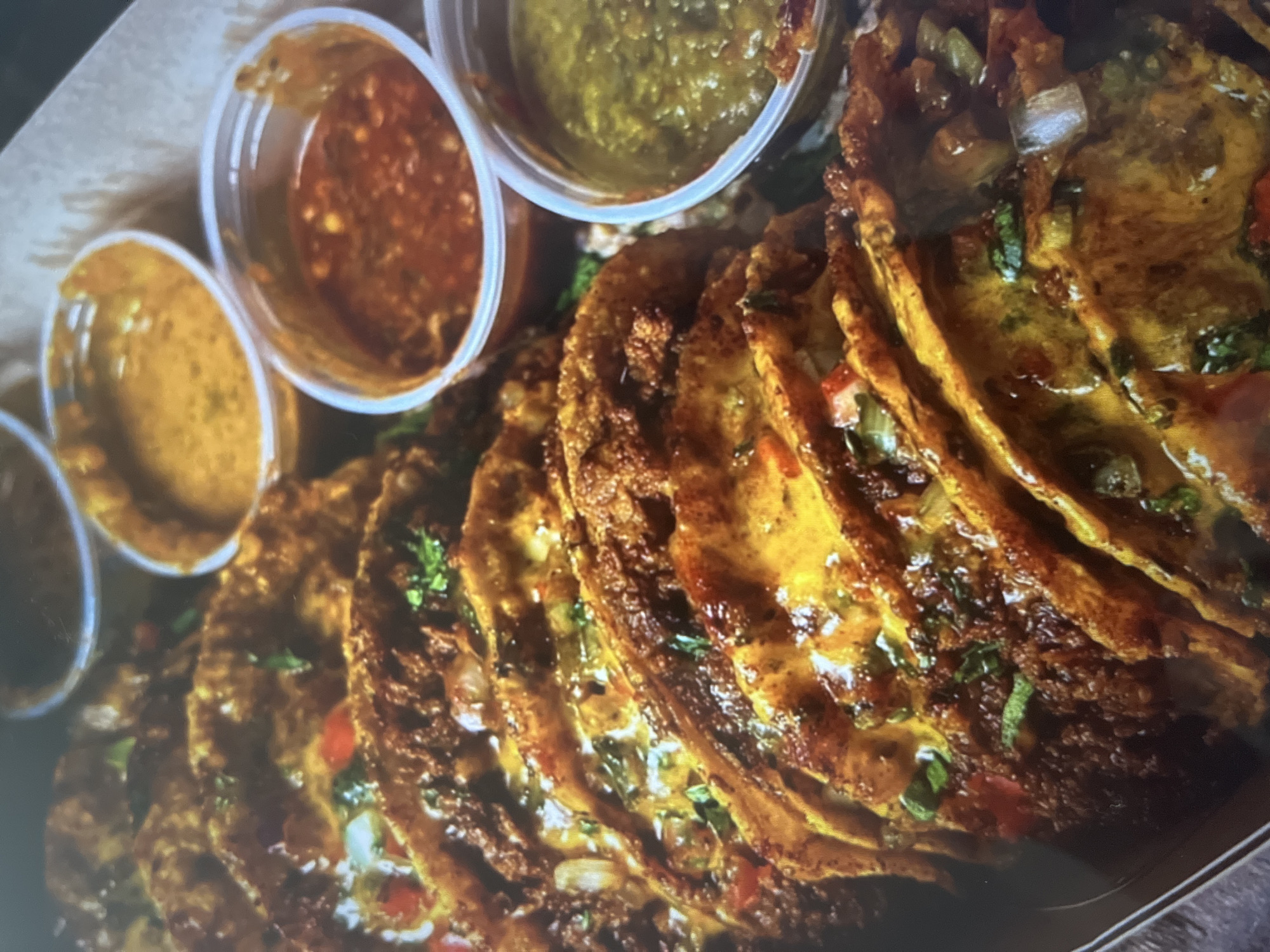 A close-up, overhead shot shows a tray of crispy tacos arranged in rows, with small plastic cups of dipping sauces placed behind them. The tacos are golden brown and appear to be filled with a dark, ground meat mixture, visible bits of red bell pepper, and white onion. Melted cheese and chopped cilantro are visible on top of the tacos. The dipping sauces are in separate cups, with one appearing to be a reddish-brown salsa, another a yellowish sauce, and a third a green salsa. The background is out of focus and appears to be a dark cardboard container. The image does not contain any information about the location in the city of Unknown, Unknown, nor does it show any people or actions.