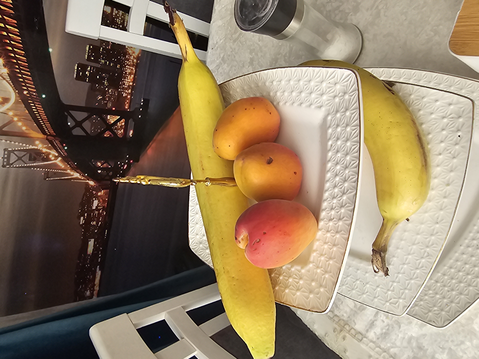 A still life composition features a fruit arrangement on a textured white serving platter, which is placed on a marble-patterned surface. The platter holds three ripe apricots and a long, yellow banana. Another banana lies horizontally on a second, larger serving platter beneath the first. Behind the fruit, a salt shaker with a black lid and a clear base is visible. In the background, a large mural or tapestry depicts a nighttime city scene with a bridge illuminated by numerous lights, suggesting an urban setting. A white chair is partially visible in the foreground and to the left. The lighting appears to be artificial, casting soft shadows and highlighting the fruits' textures and colors. The overall mood is domestic and relaxed. No visible text is present in the image.