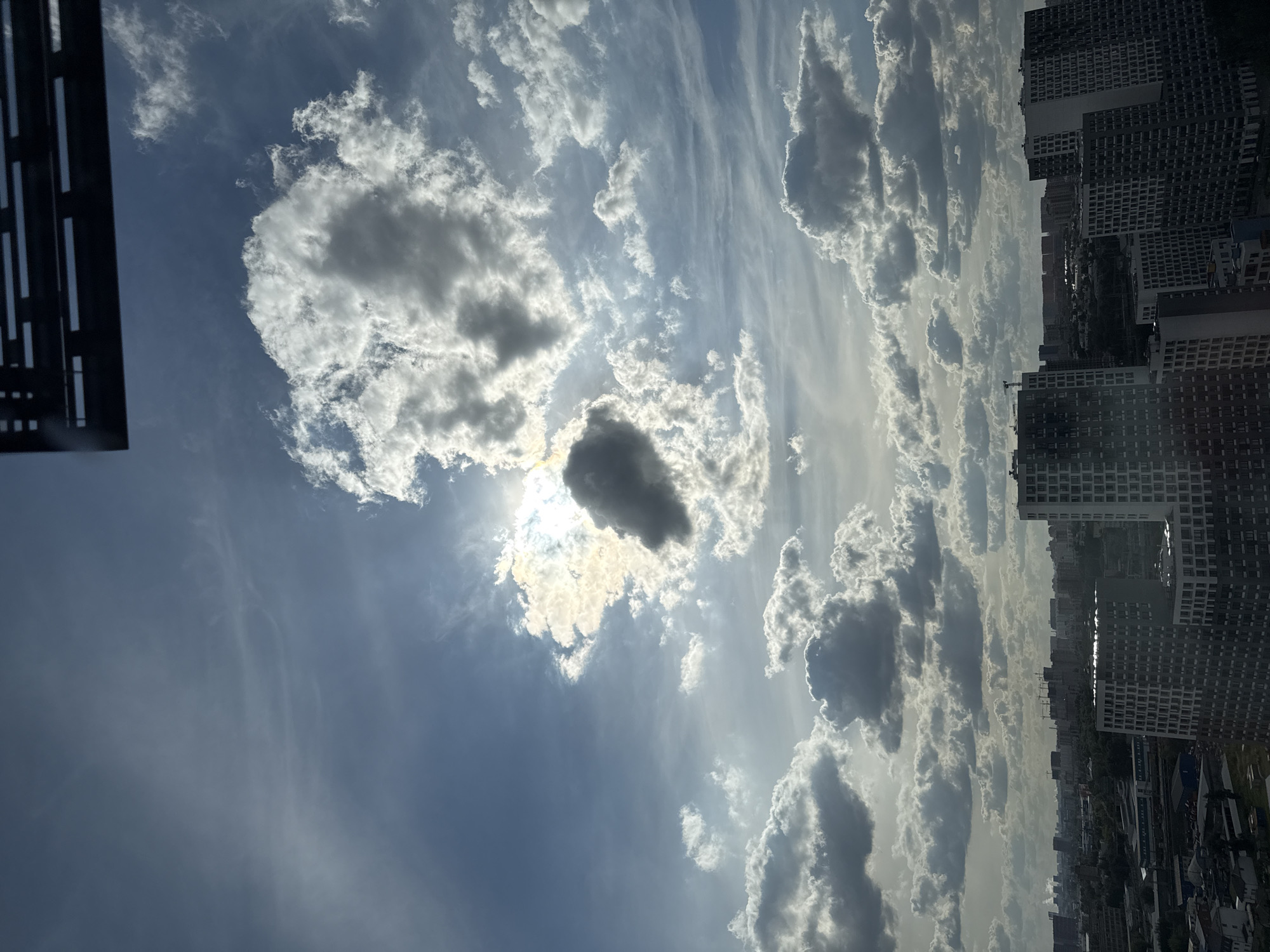 The image shows a cityscape under a partly cloudy sky. The foreground features several tall residential buildings with modern architecture. In the midground and background, more buildings and structures are visible, suggesting a dense urban environment. The sky is a mixture of blue and grey clouds, with the sun partially obscured by a large, dark cloud formation. The sun's rays are breaking through, creating a dramatic lighting effect. The scene appears to be a panoramic view of the city, possibly taken from a high vantage point. The location is the city of Lyubertsy, Russia.