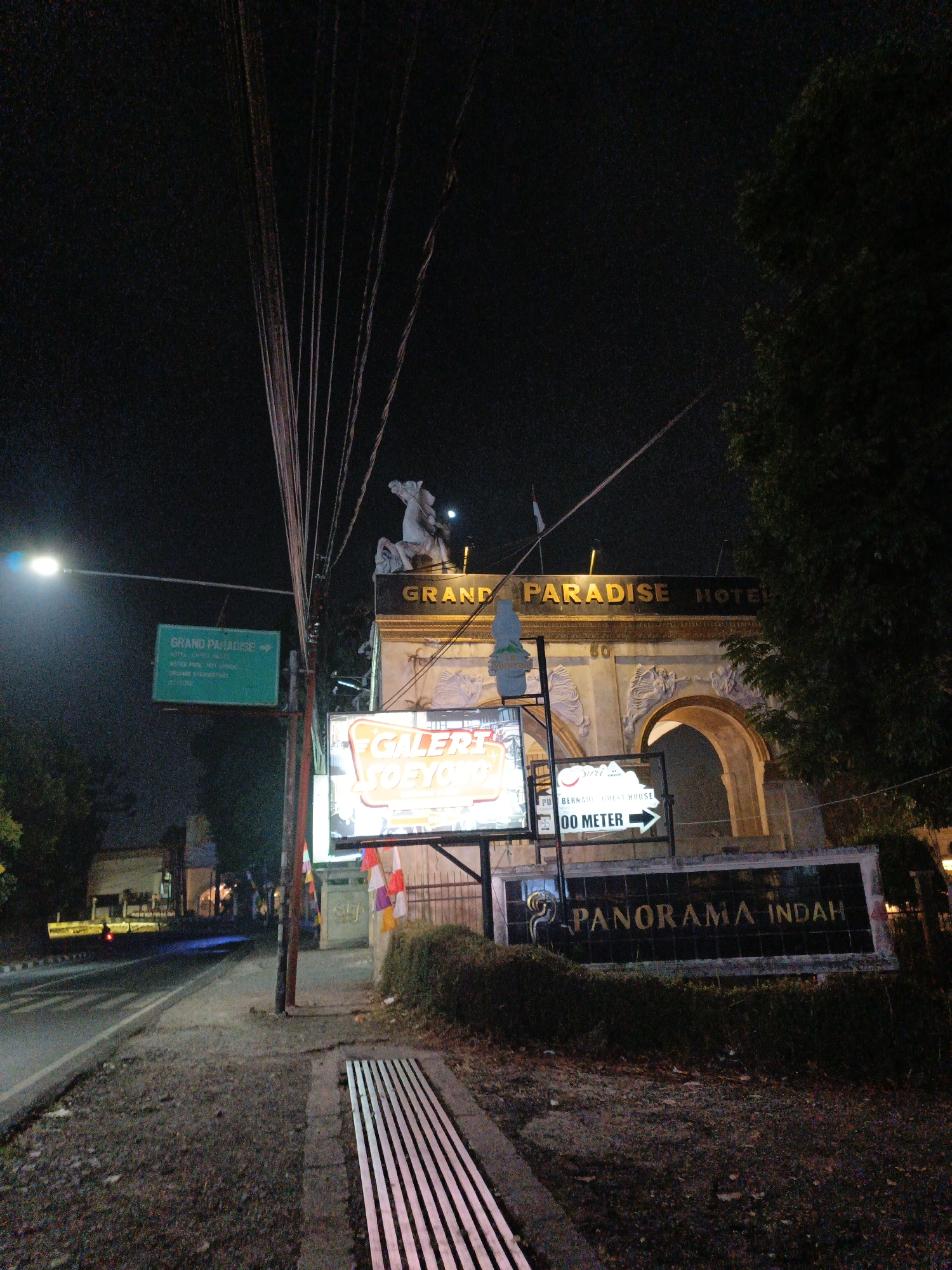 The image depicts a street scene at night in Lembang, Indonesia. In the foreground, a concrete sidewalk with a metal grate runs along the right side of the image. To the left, a road is visible with a lane marker and a passing vehicle with illuminated headlights.
Dominating the center of the image is the entrance to "Grand Paradise Hotel". The architectural structure features a large archway and is illuminated by a sign bearing the hotel's name. Above the arch, a statue of a horse and rider is visible.
Several signs are present:
- A green directional sign on the left reads "GRAND PARADISE" with an arrow pointing right and smaller text below indicating "HOTEL", "WATER PARK", and other services.
- A brightly lit neon sign reads "GALERI JOEYO".
- Another sign below the "GALERI JOEYO" sign indicates "PU BERNADI GUEST HOUSE 100 METER" with an arrow pointing right.
- A large, dark sign with illuminated text reads "PANORAMA INDAH".
Decorative flags in red and white are attached to a pole near the "GALERI JOEYO" sign. Overhead, a network of electrical wires crisscrosses the sky. The environment appears to be an urban or semi-urban area with trees and vegetation visible on the right side. The overall lighting suggests it is nighttime, with artificial light sources illuminating the signs and road.