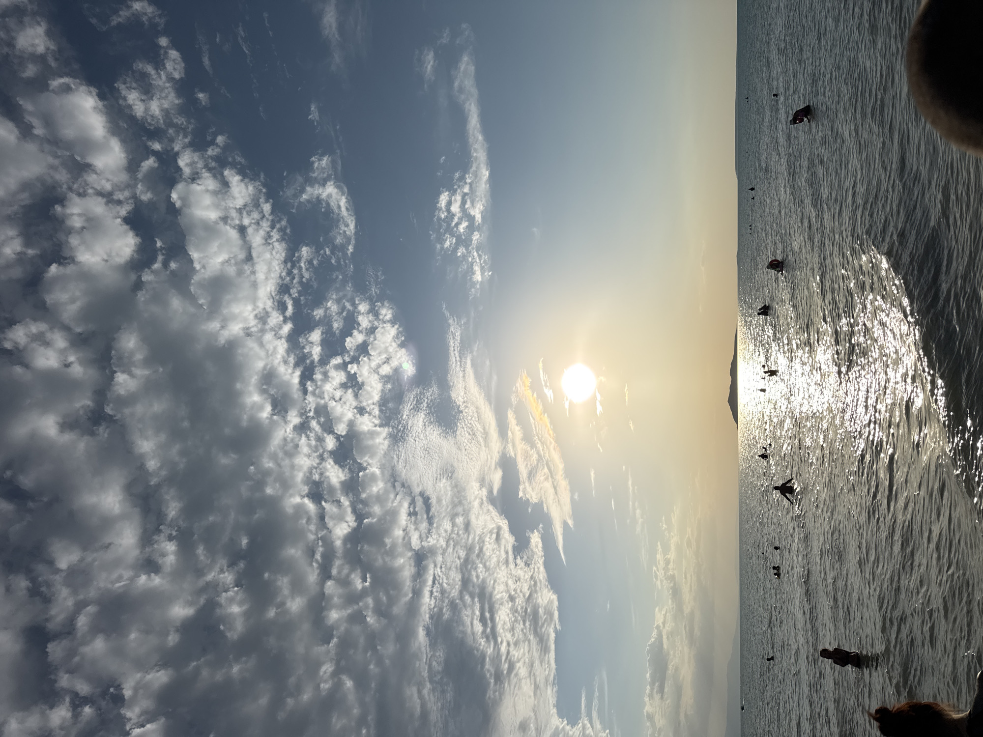 In the city of Zharbulak, Kazakhstan, a tranquil sunset scene unfolds at a beach. The sun, a bright orb, is partially obscured by scattered clouds in the sky, casting a warm, golden glow across the water. The sea is calm, with gentle ripples reflecting the sunlight, creating a shimmering effect. Several individuals are enjoying the water, some wading, others swimming, and a few are silhouetted against the setting sun. A distant island or landmass is visible on the horizon, adding depth to the landscape. The overall atmosphere is peaceful and serene, capturing a moment of relaxation by the sea.