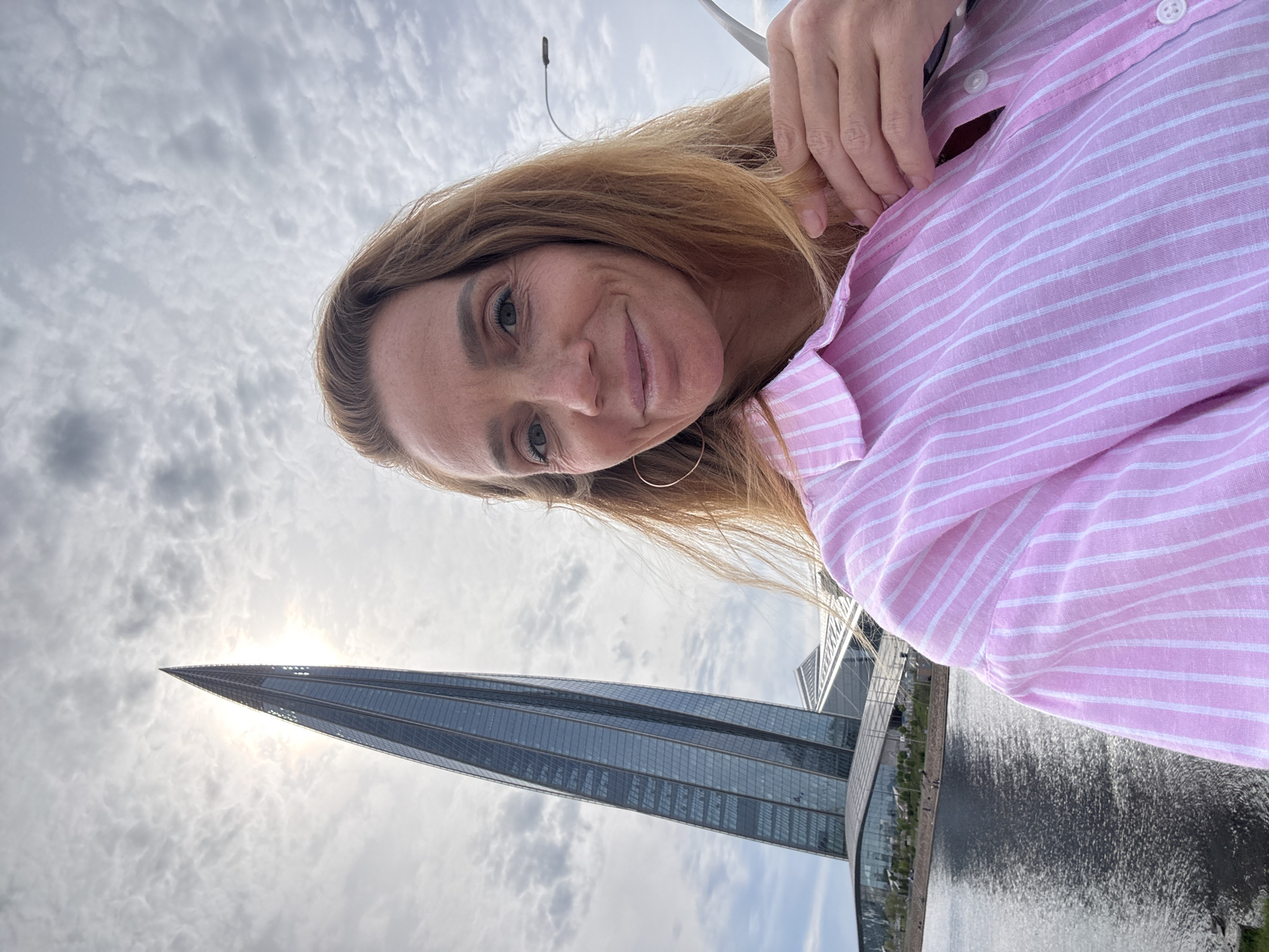 A woman is pictured outdoors, likely taking a selfie. She has long, light brown hair and is wearing a pink and white striped shirt. She is smiling at the camera.
Behind her, a modern skyscraper, likely the Lakhta Center, is visible. A body of water, possibly a river or lake, is also in the background. The sky is overcast with cloudy conditions. This scene is located in Saint Petersburg, Russia.
