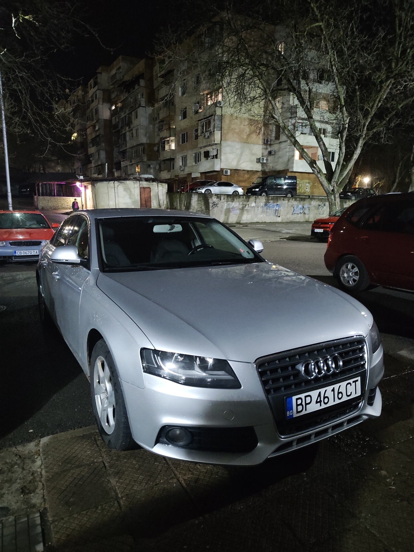 The image shows a silver Audi car parked on a street in Kozloduy, Bulgaria. The license plate number is BP 4616 CT. Behind the car, there are several parked cars, including a red hatchback and a red sedan with license plate CB 0670 TA. In the background, there is a multi-story apartment building with lights in some of the windows and bare trees in front of it. The street is dark, suggesting it is nighttime. A person is visible standing in front of a small structure.
