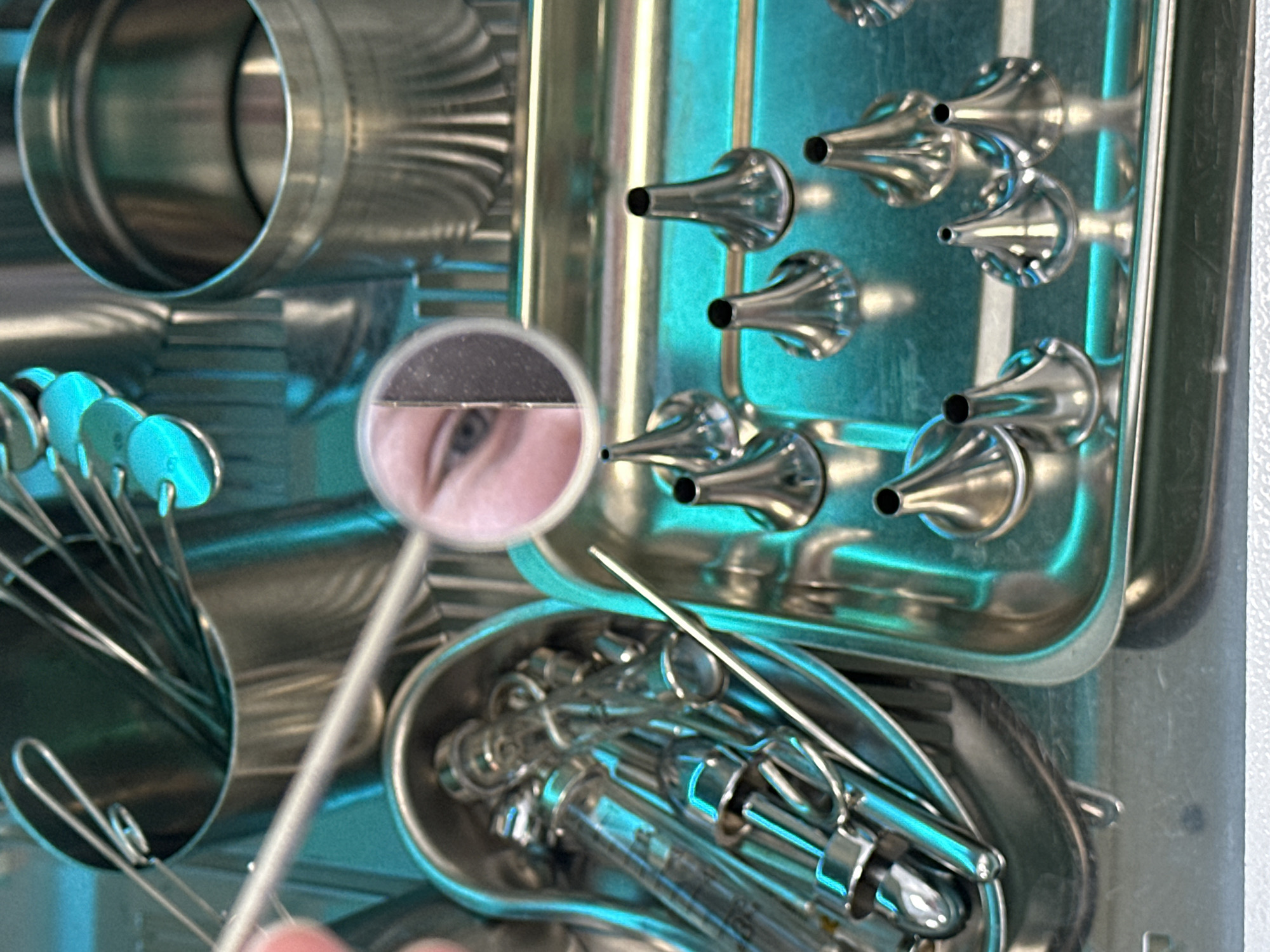 A close-up photograph shows a collection of medical instruments, including otoscopes, mirrors, and syringes, arranged in stainless steel trays and containers. The instruments are clean and appear to be sterilized, with some showing reflections of a blueish light. A round mirror held by a thin metal handle reflects a portion of a face, showing an eye and part of the forehead. The location, Kazan, Russia, is not directly evident from the image.