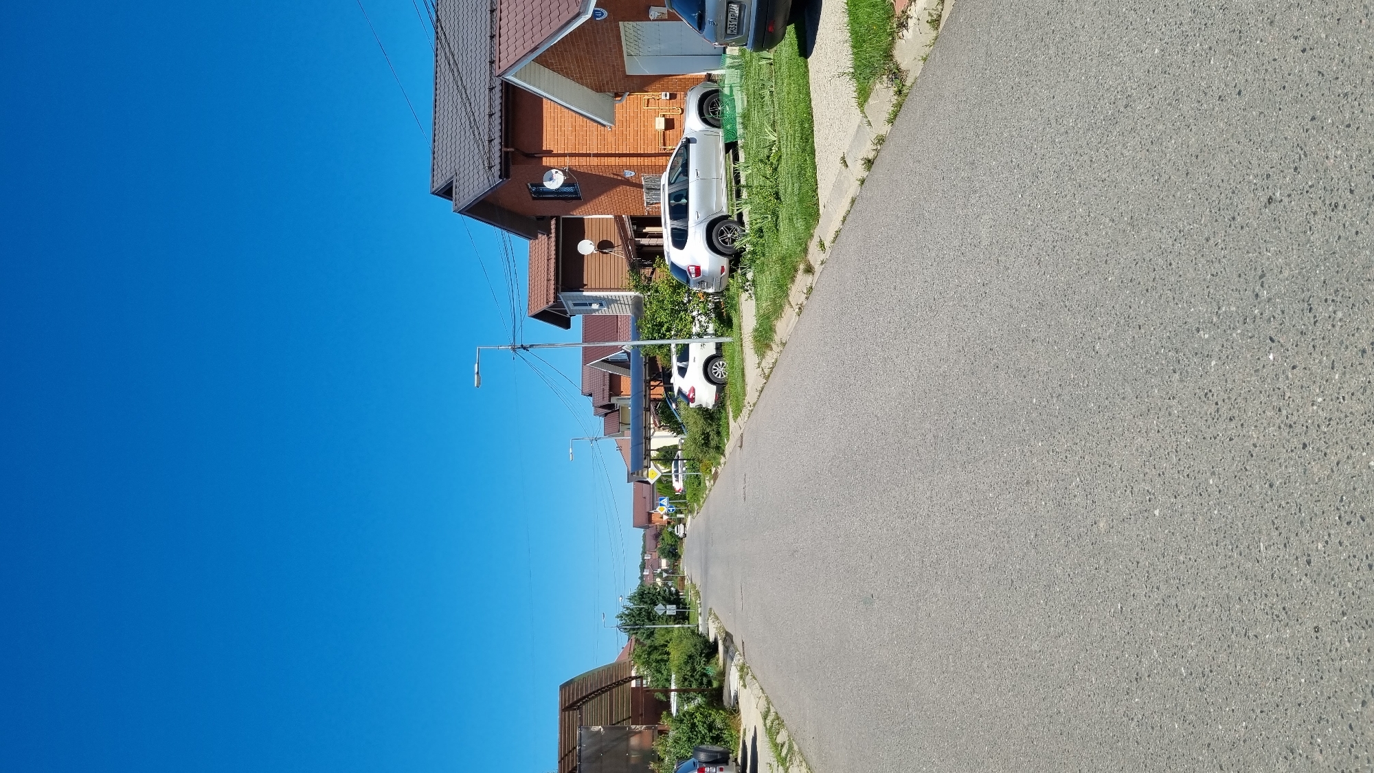 The image depicts a quiet residential street in the village of Verzilovo, Russia, on a clear, sunny day. The asphalt road runs towards the horizon, flanked by well-maintained houses with terracotta-tiled roofs. Several cars are parked along the street and on the lawns in front of the houses. These include a silver SUV, a white SUV, and the rear of a silver car. Lush green trees and bushes line the street, interspersed with streetlights and power lines. The sky is a vibrant, cloudless blue. The scene conveys a sense of peaceful suburban life.