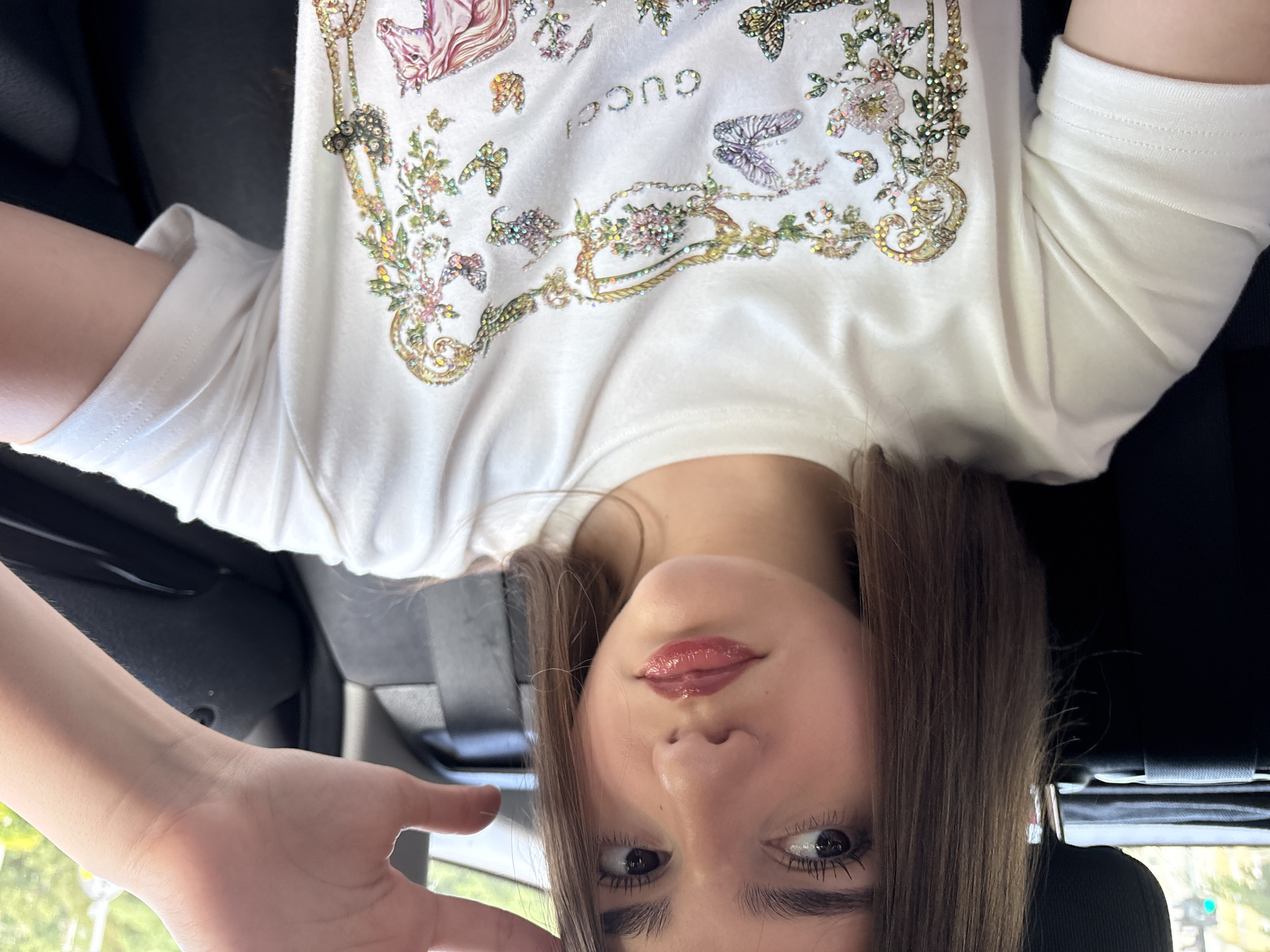 A young female is seated within the passenger compartment of a vehicle. Her straight, brown hair frames a light-toned face. She possesses dark eyes with emphasized eyelashes and full, glossy lips with a pinkish tint. Her right hand is partially visible in the upper right quadrant of the image, with fingers slightly bent. She is wearing a white short-sleeved t-shirt featuring an elaborate, multicolored, glitter-embellished design. This design incorporates golden scrollwork, clusters of small flowers, multiple iridescent butterflies, the word "GUCCI" in similar iridescent letters, and a partial depiction of a pink and purple unicorn or horse. The interior of the vehicle shows dark upholstery on a headrest and seat, and a dark door panel. Through the car window, a blurred outdoor environment is visible, characterized by green foliage and a partial green light, suggesting a street scene in an unknown city. The individual is looking directly towards the camera.