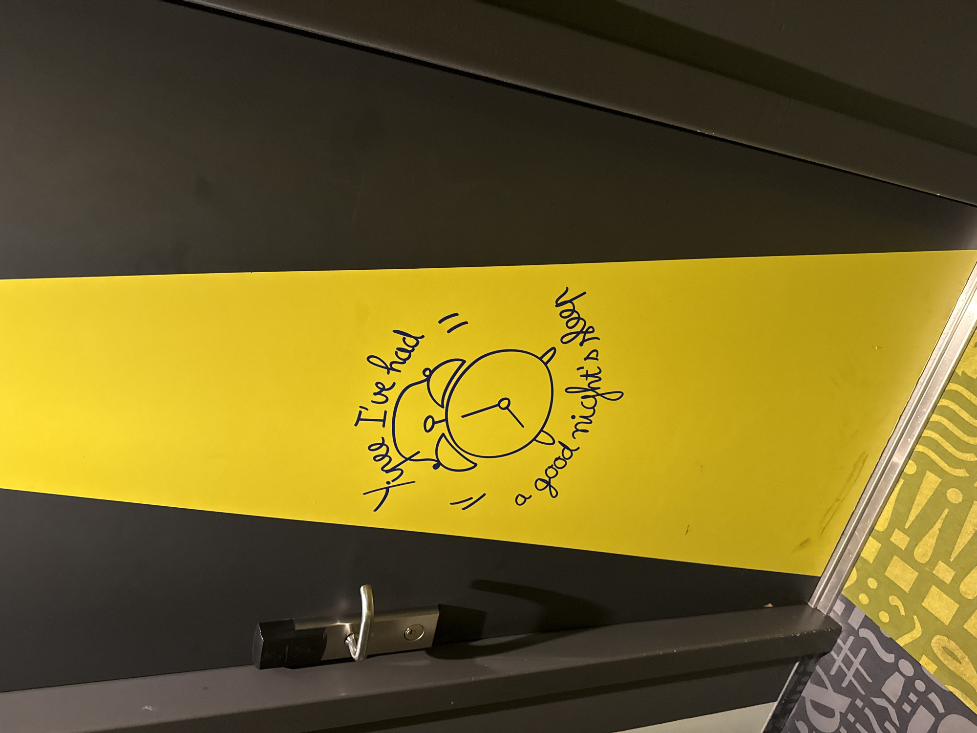 The image depicts a close-up, eye-level, slightly angled view of a dark gray door with a bright yellow vertical stripe. On the yellow stripe, a drawing of an alarm clock and handwritten text are present. The text reads "Yipee I've had" with quotation marks on the right, followed by "a good night's sleep" below. The drawing of the alarm clock is in dark blue ink, as is the text. To the left of the yellow stripe, a silver door handle and lock mechanism are visible on the dark gray door. Below the door, a portion of the floor is visible, with a gray carpet on the left featuring a repeating pattern of symbols like "#" and "A", and a yellow mat on the right with a repeating pattern of dots, commas, exclamation points, and wavy lines. The scene appears to be indoors, likely a hallway, and given the context provided about Stein, Netherlands, it's possible this is a hotel or a residential building. The image quality is good, with clear focus on the door and its decorations.