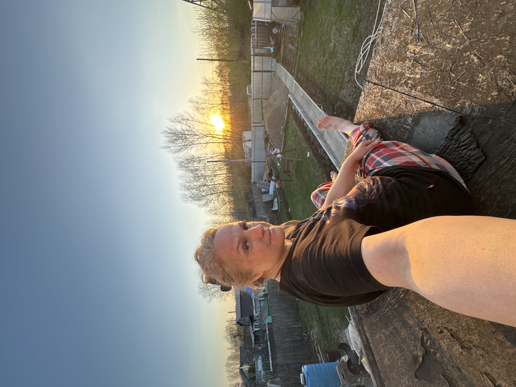 A person sits on a flat, weathered, rough surface, possibly a rooftop, overlooking a landscape featuring a sunset. The person is wearing a black t-shirt, red and blue plaid pants, and is barefoot. The landscape includes a garden area, a fence, trees, and buildings in the distance, bathed in the warm glow of the setting sun. The location is likely in a rural or suburban area within the city of Chaikovsky, Russia, given the typical residential and agricultural structures present. A section of white rope is visible on the rooftop.