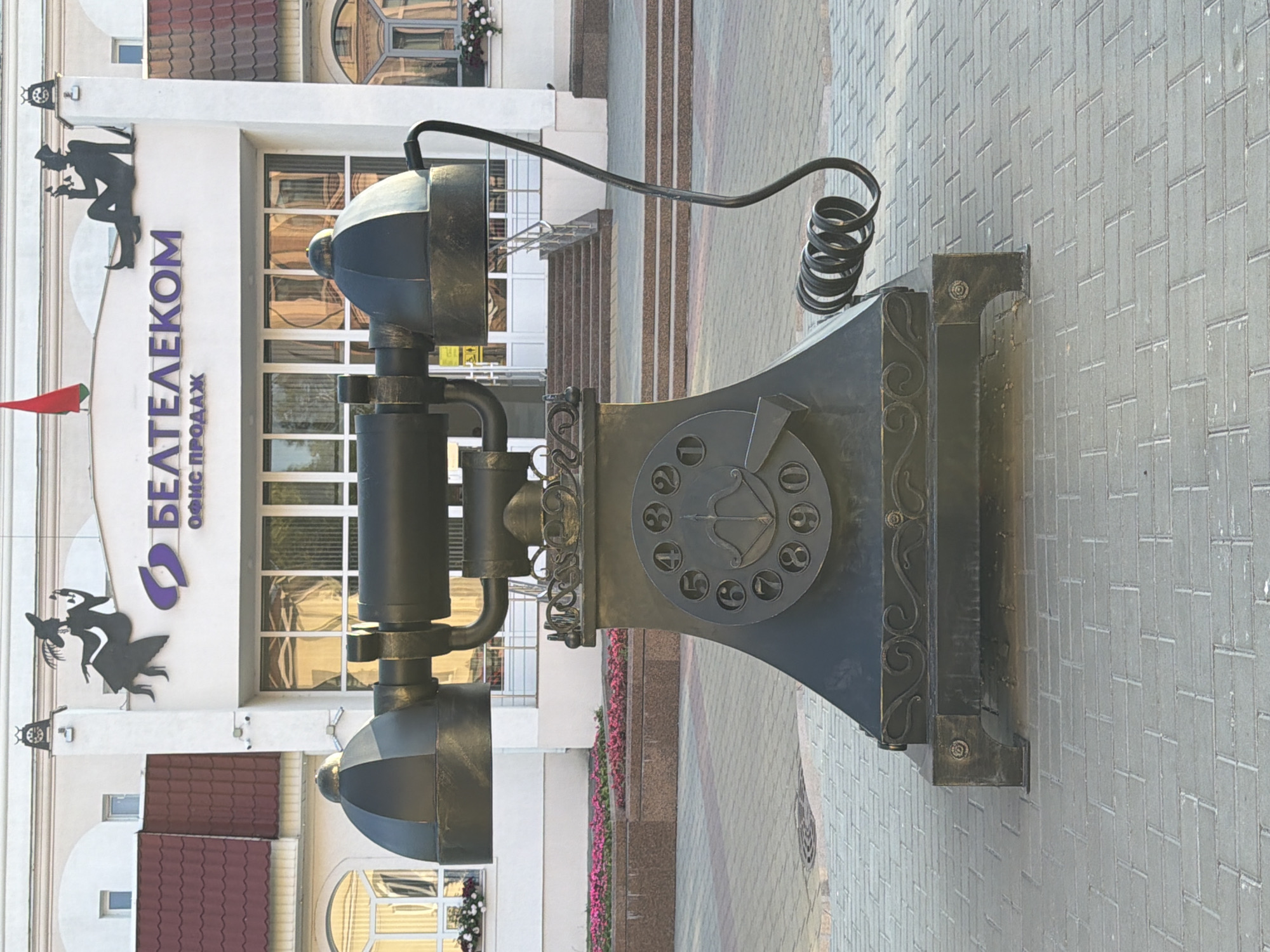 A large, dark-colored, metal sculpture of an old-fashioned telephone stands vertically on a sidewalk. The sculpture features a receiver with a coiled cord, a dial with numbers, and decorative scrollwork on the base. The background shows the facade of a modern building with large windows and a prominent sign that reads "БЕЛТЕЛЕКОМ" in purple letters, with smaller text below it. There are also decorative silhouetted figures of people on the building. The scene appears to be outdoors, possibly in front of an office building or business. The sidewalk is paved with grey bricks. There is no indication of the time of day or weather conditions.
