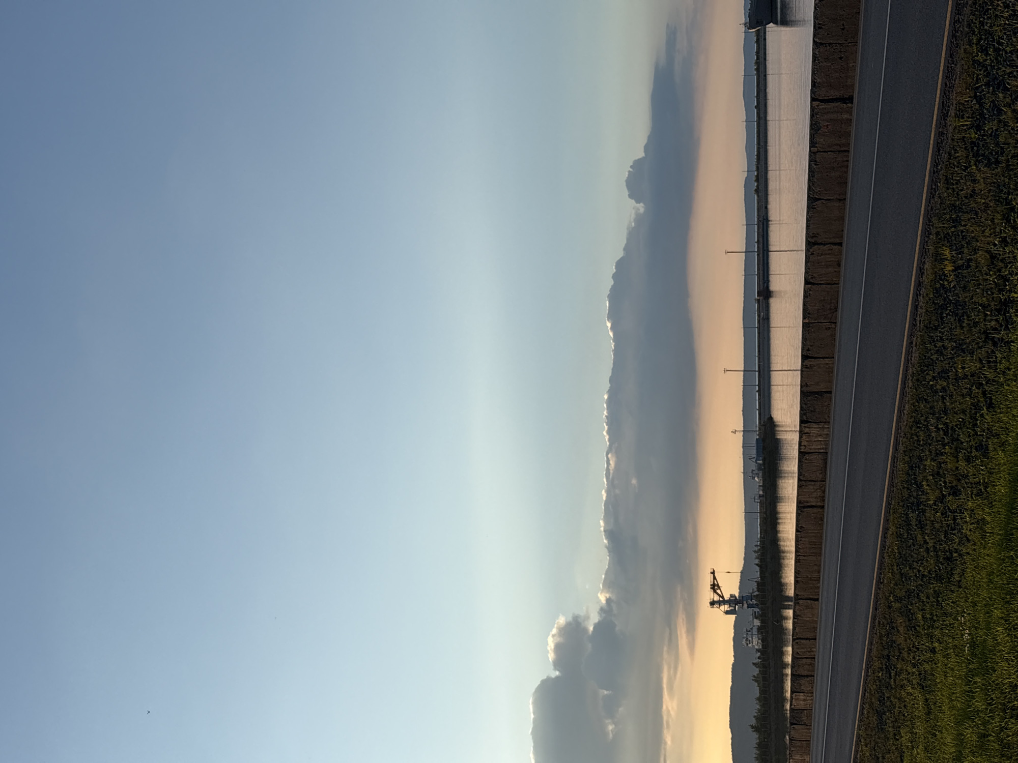The image captures a serene landscape scene, likely in Chaikovsky, Russia, during what appears to be late afternoon or early evening. The sky is a gradient of soft blues and yellows, with hints of warm light filtering through the clouds, suggesting a sunset or sunrise. A large, imposing cloud formation stretches across the horizon, its edges illuminated by the setting sun.
In the foreground, a paved road runs diagonally across the frame, bordered by a strip of lush green grass on the right. The road is marked with white lines, indicating a clear path. To the left of the road, a body of calm water reflects the muted colors of the sky and the distant landscape. The water is bordered by a series of upright posts, possibly part of a fence or structure.
Further in the distance, a prominent silhouette of a large industrial crane is visible against the horizon. It stands out due to its skeletal structure and its placement within the softer, more muted tones of the background. Several other smaller structures or industrial elements can be seen in the vicinity of the crane. The overall mood of the image is tranquil and expansive, with a sense of open space and quietude. There is no visible text in the image.