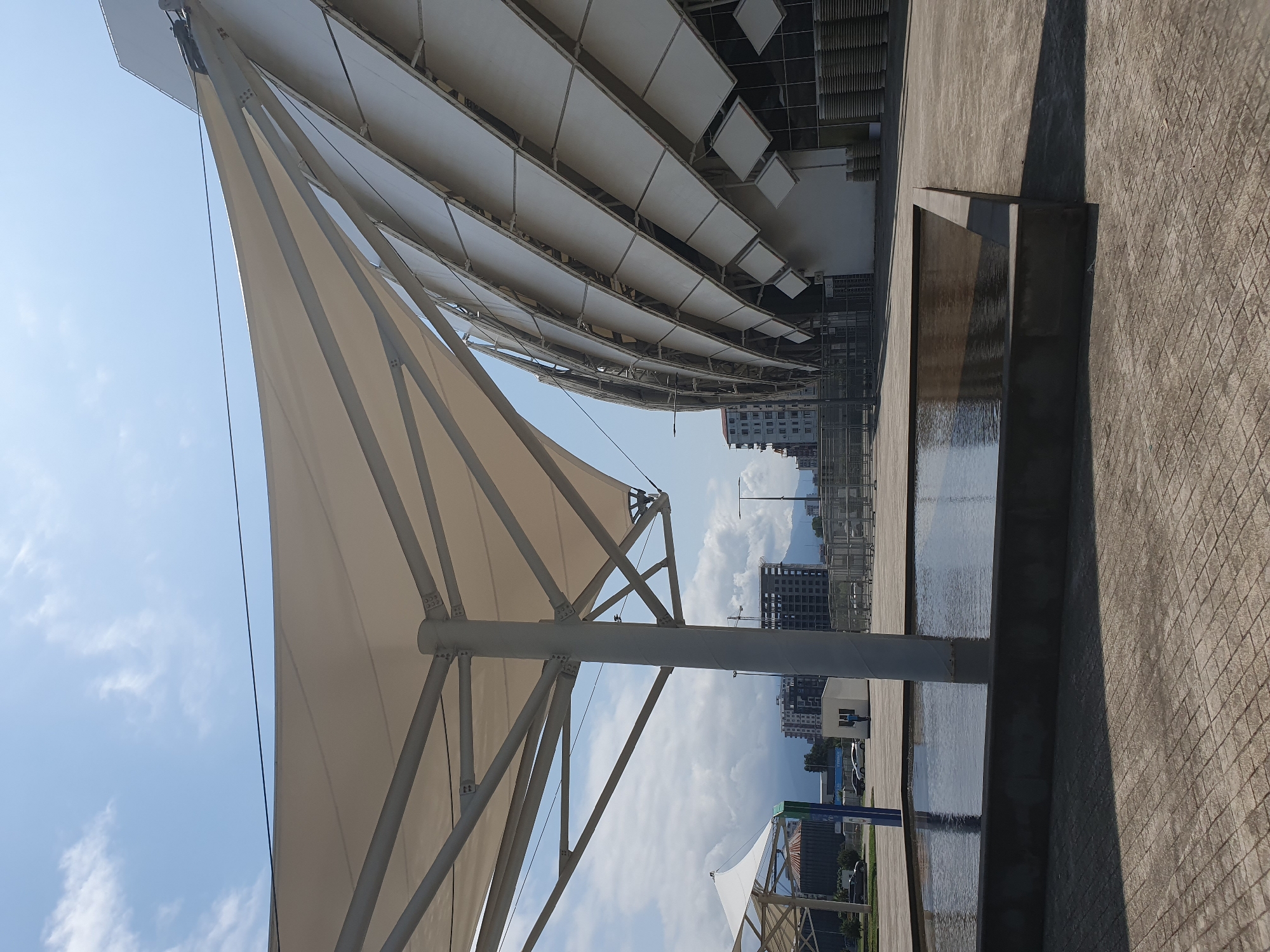The image shows a modern architectural structure in Batumi, Georgia, likely a stadium or arena. In the foreground, a shallow water feature with dark grey edging is visible, reflecting the sky and overhead canopy. The paving stones around the water feature are a light grey, laid in a herringbone pattern. A large, white tensile fabric canopy supported by a central grey pole dominates the left side of the frame. Behind the canopy, a portion of a curved building facade made of white panels and metal supports is visible, suggesting a large, contemporary facility. In the background, several multi-story buildings, some appearing under construction, are present. A clear blue sky with scattered white clouds is overhead. A few cars are parked in the distance. No people are clearly discernible in the scene. The overall impression is one of a modern, open public space with striking architectural elements.