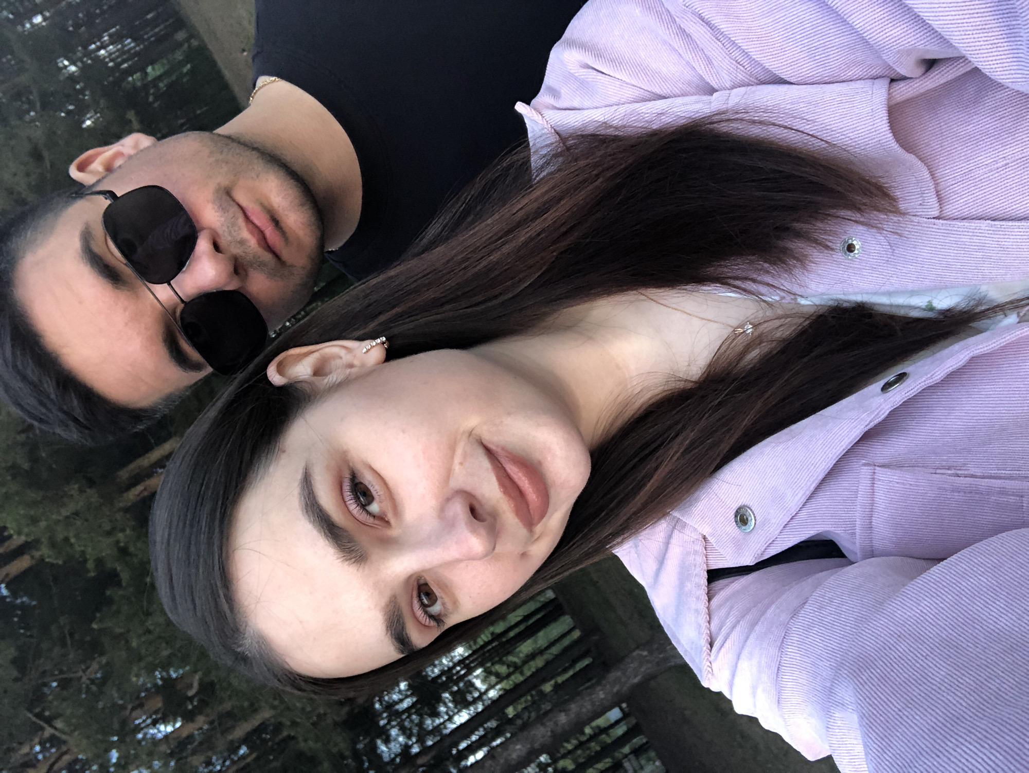 A selfie shows two people, possibly a couple, in an outdoor setting. The woman, with brown hair and wearing a light purple jacket, is closer to the camera and angled in the shot. The man, wearing sunglasses and a black shirt, is slightly behind her to the left. The background appears to be a wooded area with trees. Given the prompt includes the city of Chaikovsky, Russia, the setting is likely within or near that city.