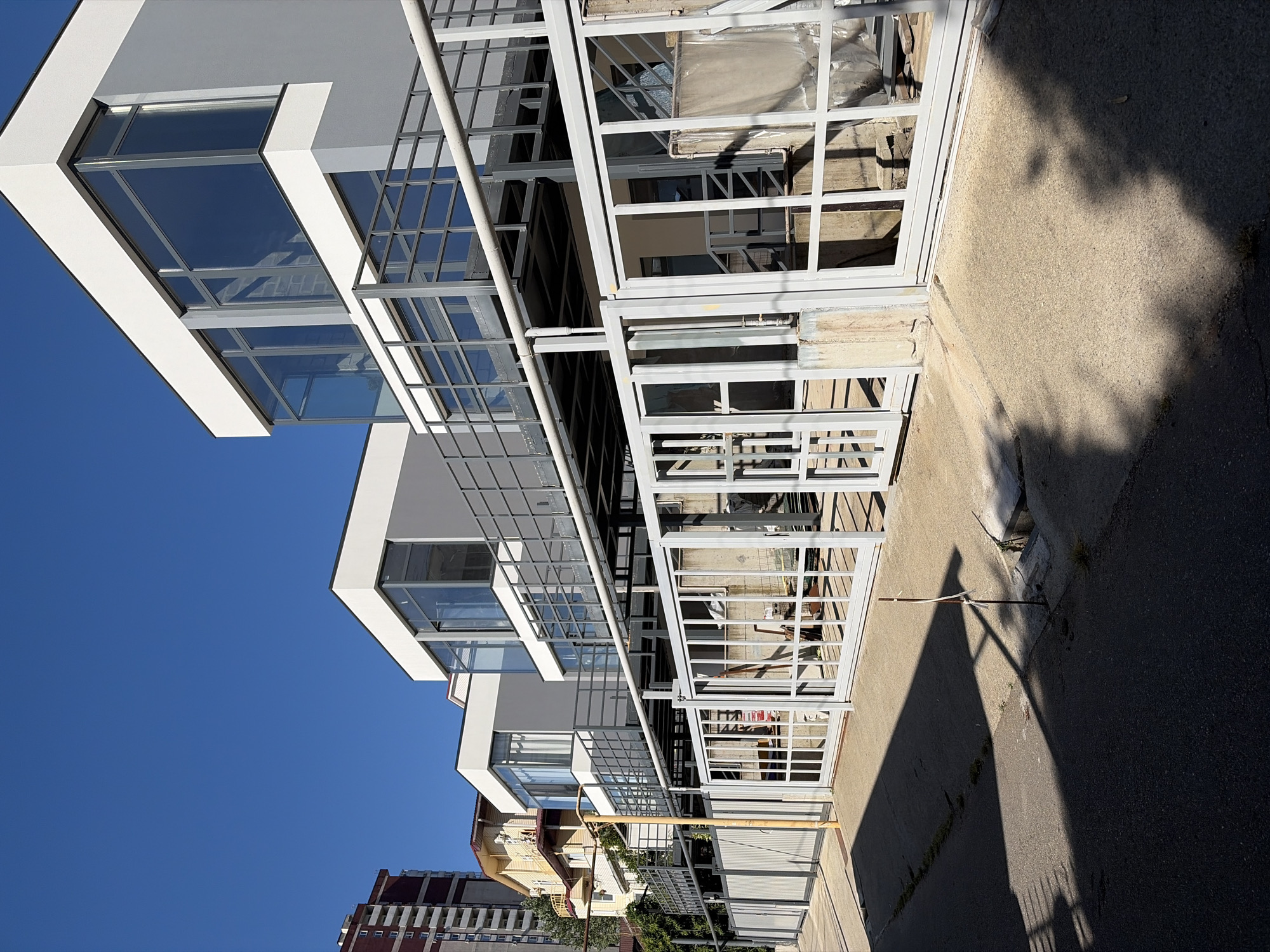 The image depicts a row of modern, two-story residential buildings with large glass windows and grey and white exteriors, set against a clear blue sky. The buildings feature balconies or elevated walkways with grey metal railings. In the foreground, a grey metal fence runs along the property line, separating it from a paved area and a street. Some vegetation is visible behind the fence. To the left, a tall, older apartment building with a reddish-brown facade stands in the background. The overall scene suggests a residential area in a city, possibly Sochi, Russia, characterized by a mix of contemporary and older architecture. There are no people visible in the image.