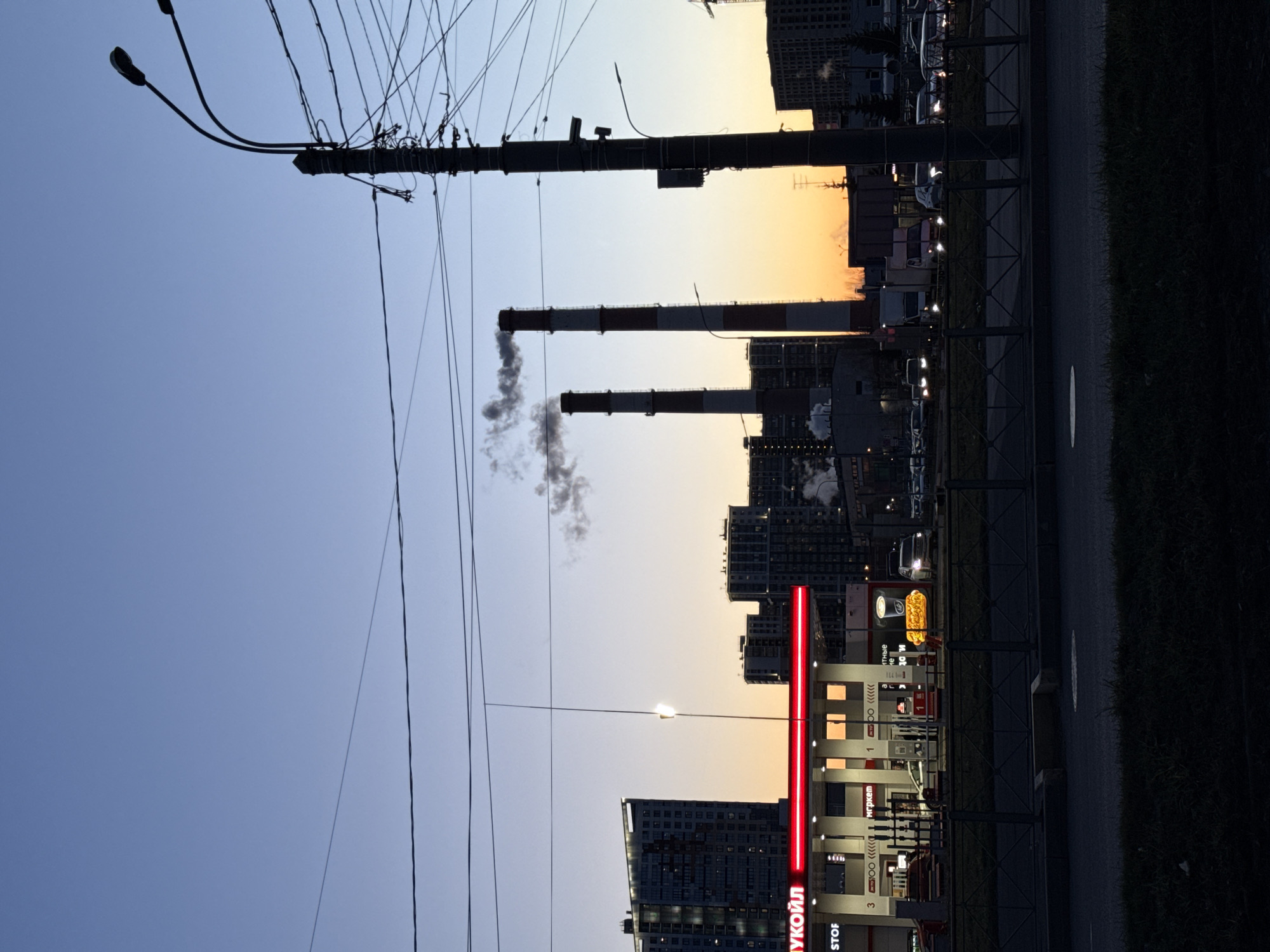 The image depicts an urban scene, possibly at dusk, within the city of Saint Petersburg, Russia. In the foreground, the red signage of a "ЛУКОЙЛ" (Lukoil) gas station is visible. A street and sidewalk run parallel to the gas station. Beyond the gas station, a series of buildings rises, with smoke stacks emitting smoke into the sky. The sky transitions from blue to orange, suggesting a sunset. Electrical poles and wires are prominent in the upper-left quadrant.