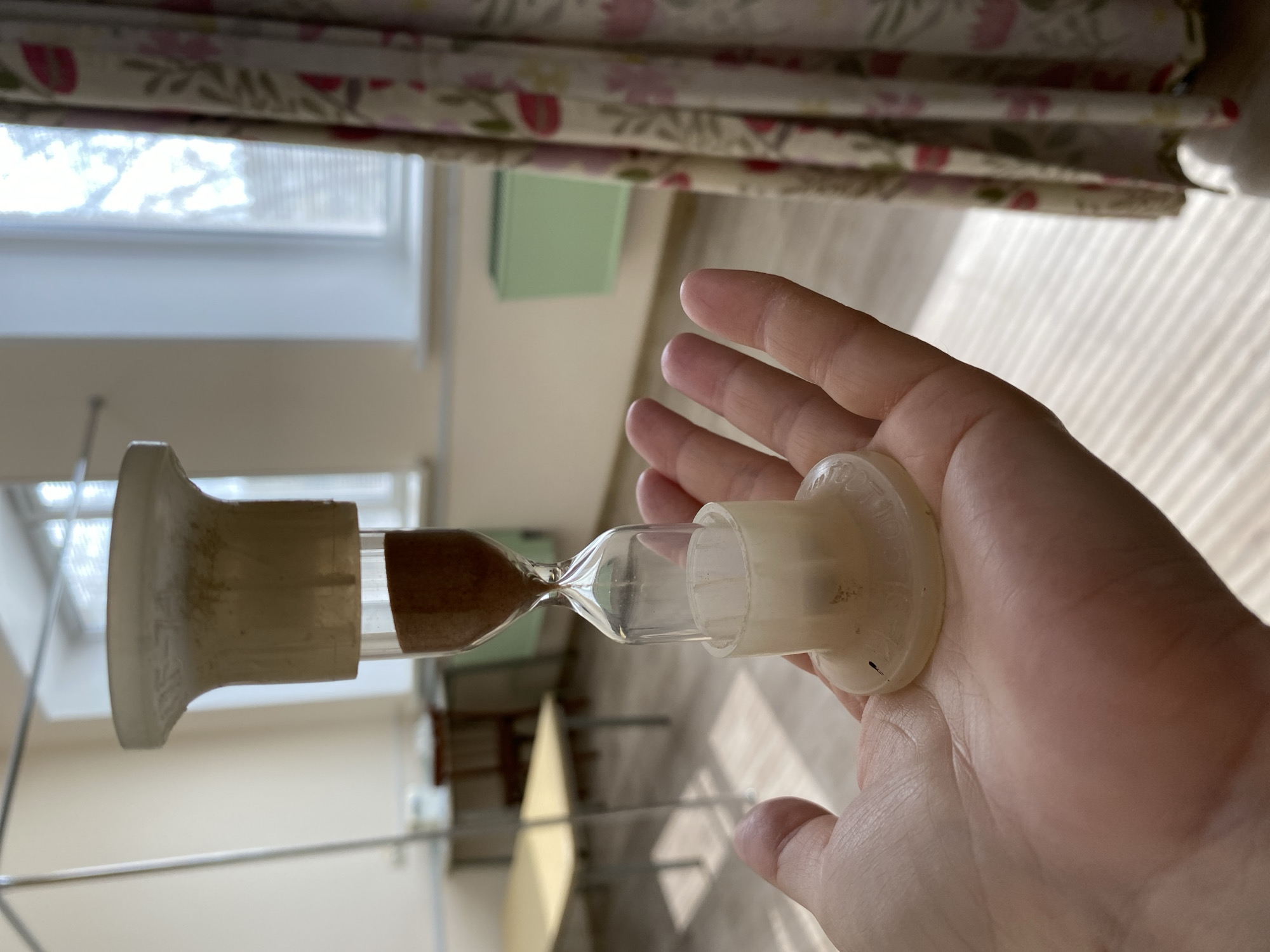 A person is holding an hourglass in their hand. The hourglass is partially filled with brown sand. The background is a domestic interior, including a window, curtains with a floral pattern, and a green object that appears to be on a wall or shelf. The photo seems to be taken indoors with natural light. It is not possible to determine the location within the city of Noviy, Russia from the image.