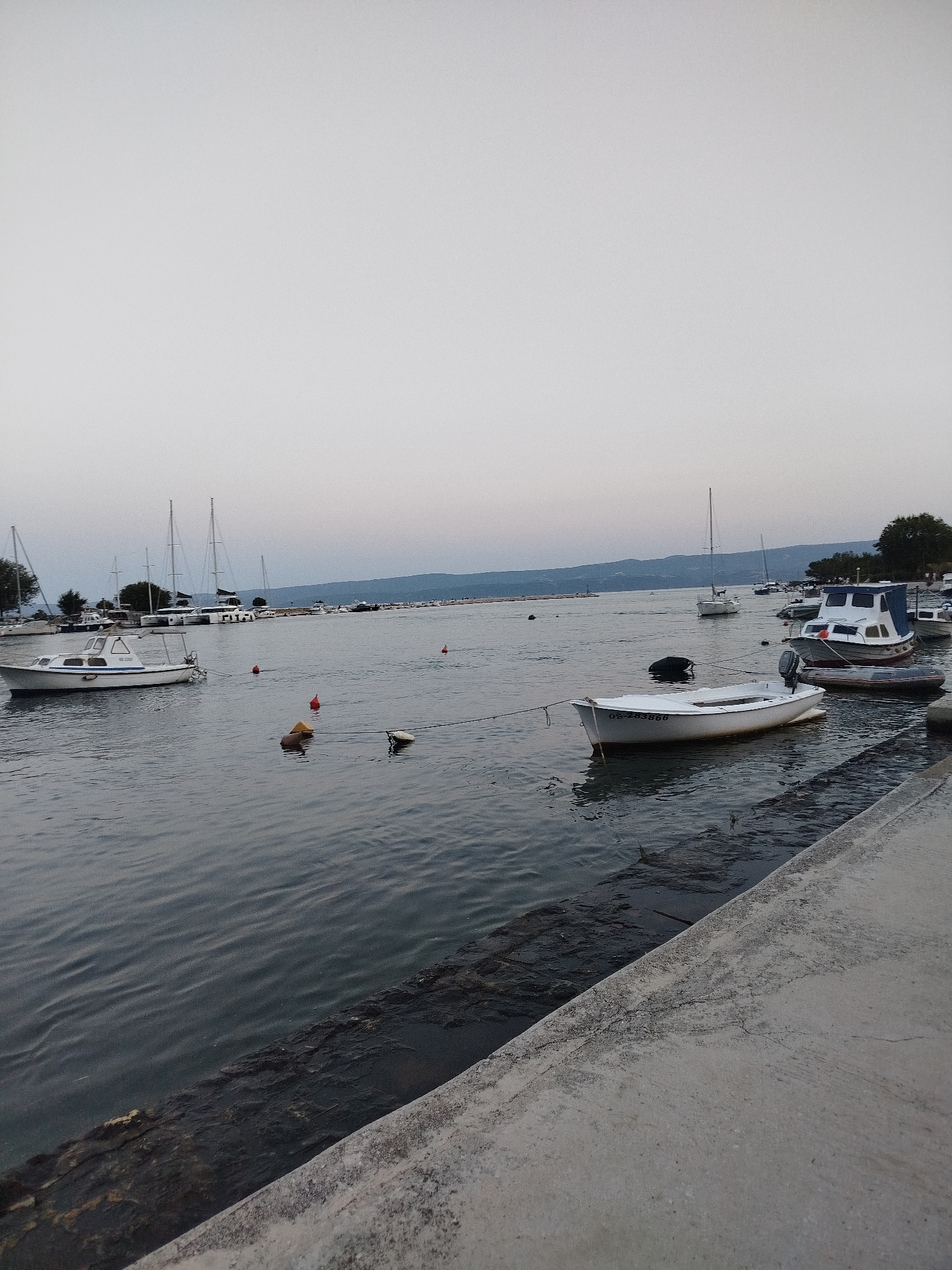 The image depicts a harbor in Omiš, Croatia, likely during the early morning or late evening due to the soft, diffused light and the muted colors. The foreground features a concrete walkway bordering the water. The water itself is calm, with gentle ripples, and several boats are moored. These include a small white motorboat with an outboard motor in the middle ground, and other vessels, including sailboats and a larger white motorboat with a blue canopy, further in the distance. Red and yellow buoys are visible in the water, marking the harbor. The background shows a coastline with trees and hills, with a hazy sky overhead. No people are visible in the scene.