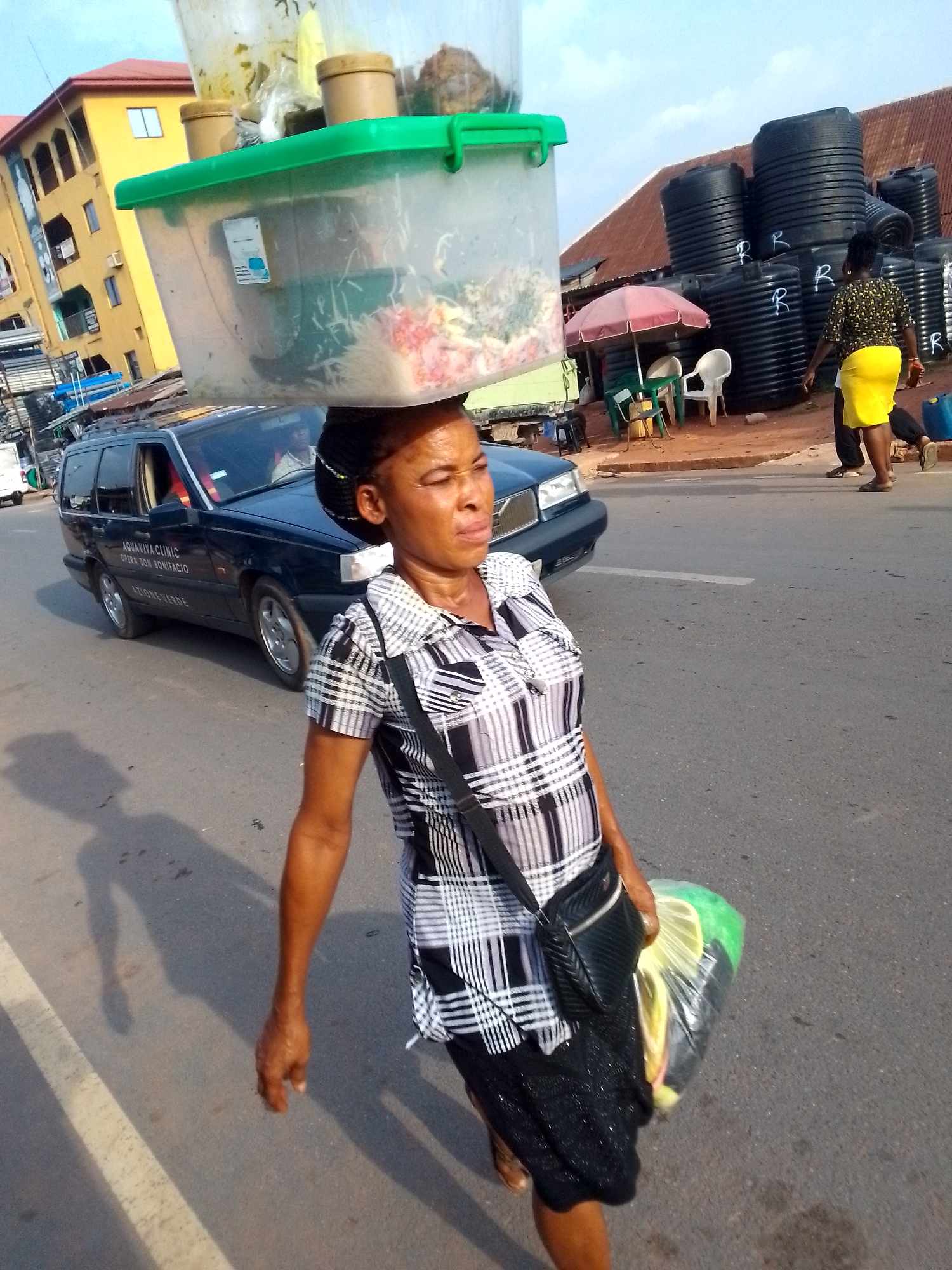 In the city of Orlu, Nigeria, a woman walks along the side of a paved road. She carries a clear plastic container, filled with various items, balanced on her head. She also carries a plastic bag filled with yellow and green objects in her left hand. She is wearing a black and white plaid shirt, a dark skirt, and a shoulder bag. A black Volvo car with writing on the side is parked behind her. In the background, there is a yellow building and a group of black water tanks stacked near a red umbrella and chairs. Another woman wearing a yellow skirt is walking away in the same direction.