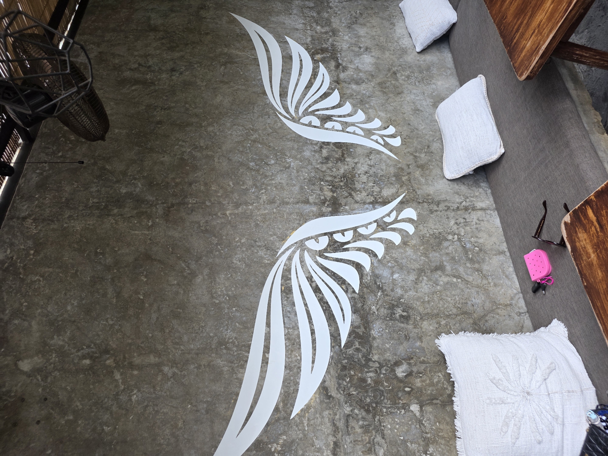 A concrete wall with a large white abstract wing mural is the central focus in this outdoor seating area in North Kuta, Indonesia. The mural is symmetrically placed, with each wing extending outwards. Below the mural, a long, gray upholstered bench is adorned with three white throw pillows, two on the right and one larger one with a textured floral design on the left. A small, bright pink pouch with a keychain is placed on the bench, along with a pair of sunglasses. To the right of the bench, a rustic wooden table with a live edge is partially visible. In the upper right corner, a dark, industrial-style ceiling fan is mounted to a slatted wooden ceiling. The overall ambiance suggests a relaxed, open-air cafe or lounge.