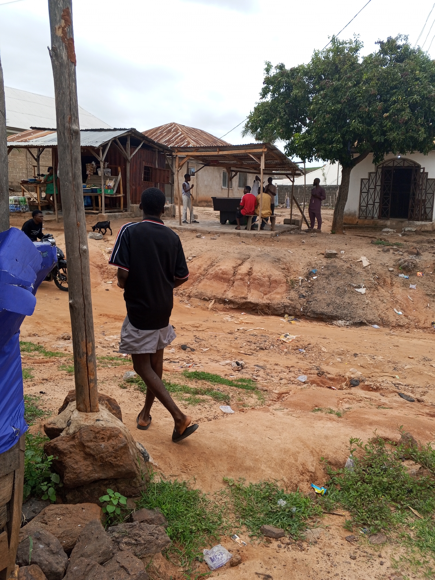 The image depicts a dusty street scene in Lokoja, Nigeria. In the foreground, a young man wearing a black polo shirt with red and white trim and grey shorts is walking away from the camera, his legs visible. To his left, a large wooden post stands next to a pile of rocks and some green vegetation. Behind him, a black goat is visible in the middle ground. Further back, there are several structures with corrugated iron roofs, appearing to be market stalls or small shops. People are gathered under an awning, with some sitting on a bench. One individual in a white shirt is holding a broom. To the right, a white building with a decorative metal-grated entrance is partially visible, with a large tree beside it. The ground is predominantly red earth and sand, with scattered debris and patches of grass. The sky is overcast.