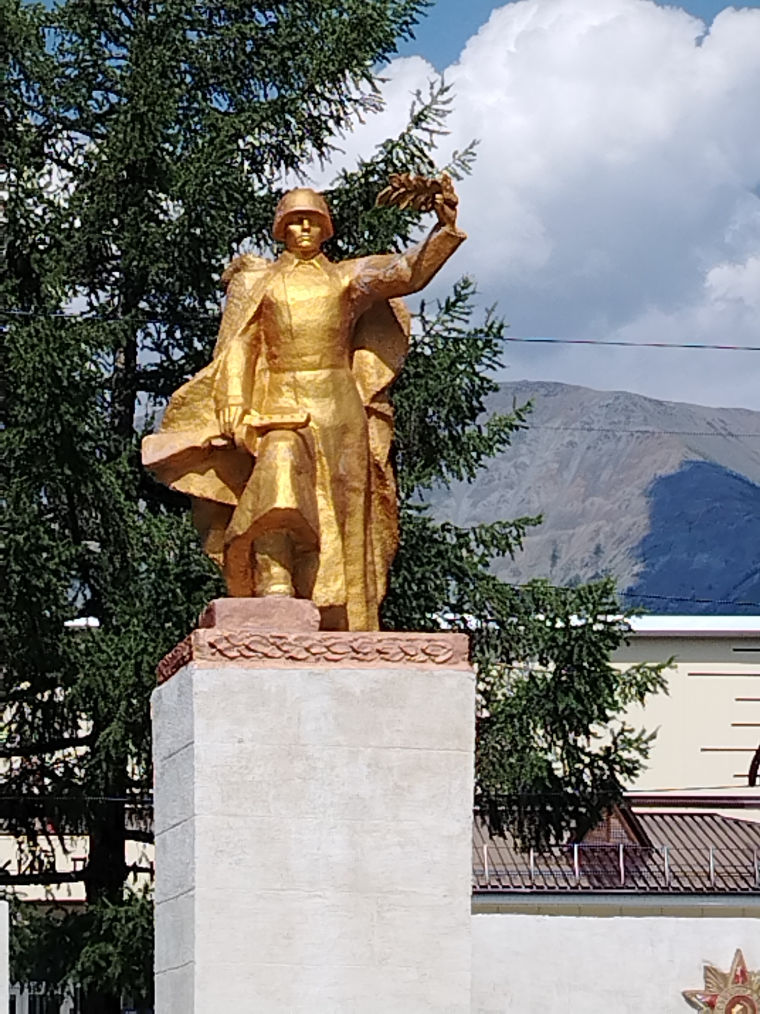 This is a close-up, waist-up, eye-level shot of a golden statue of a soldier, likely in the city of Aktash, Russia, holding a laurel wreath in his raised right hand and a sword at his side. The soldier wears a helmet and a cape that billows behind him. The statue is mounted on a light-colored stone pedestal that features a decorative, textured top edge. Behind the statue, a large evergreen tree with dense foliage dominates the left side of the frame. The background includes a muted, hazy mountain range under a cloudy sky, with a portion of a building visible on the right. The overall impression is of a monument outdoors on a bright, possibly sunny day.