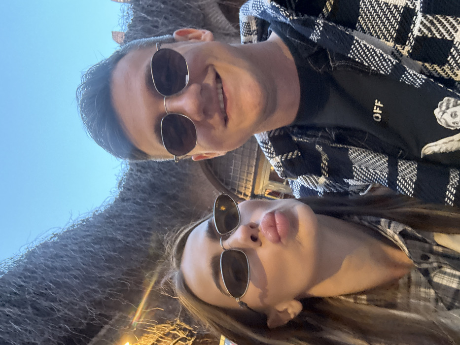 A selfie-style photograph captures two individuals, a female and a male, positioned in the foreground. The female, on the left, has long brown hair and wears a light-colored plaid shirt with oval, silver-framed sunglasses, forming a puckered lip expression. The male, on the right, is smiling and wears a black t-shirt with the partial word "OFF" visible on the chest, layered under a black and white plaid jacket, along with roundish, silver-framed sunglasses. The background features a dense, overhead canopy of bare, interwoven vines or branches. Behind the subjects, a stone archway of ancient Roman construction is visible, identifying the location as the Ponte Pietra bridge in Verona, Italy. The sky above is clear blue, suggesting daytime conditions, with a subtle light source or lens flare evident on the left side of the frame.